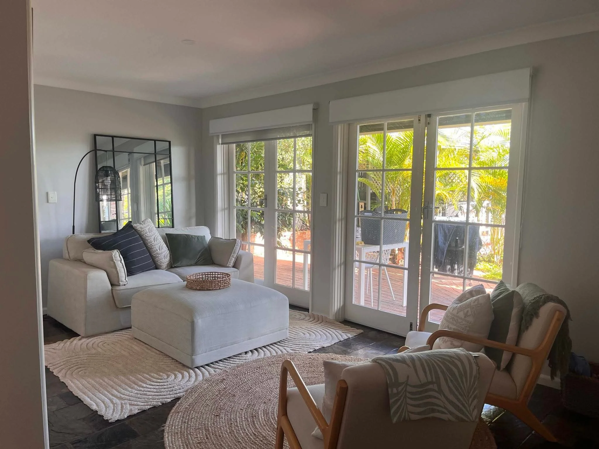 Living room with sliding glass doors opening to a patio with greenery outside. There's a white sofa with various pillows, a mirrored black frame, a floor lamp, and chairs with patterned cushions.