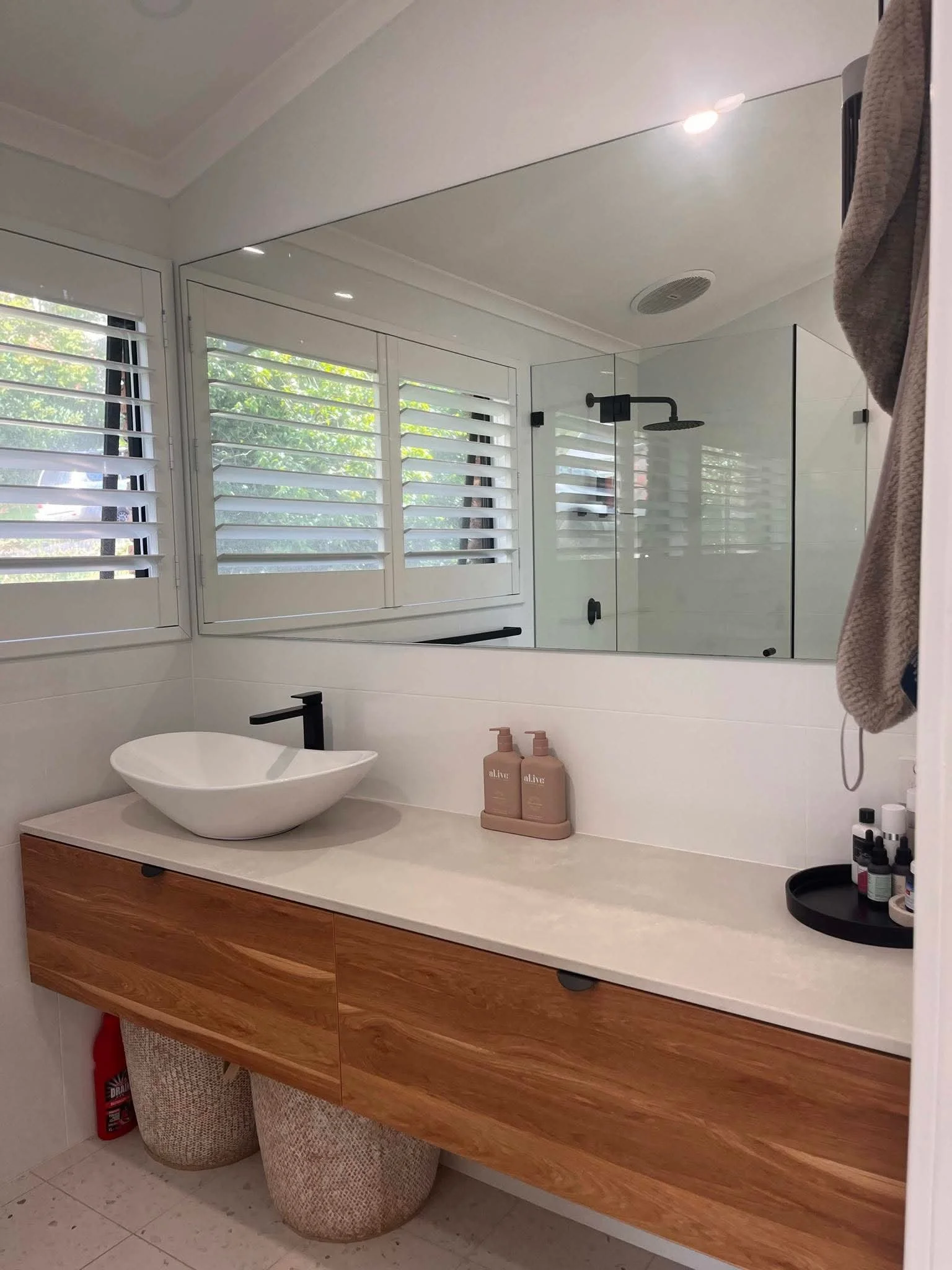 Modern bathroom with white walls and window with plantation shutters. Features a floating wooden vanity with a vessel sink, black faucet, and various toiletries. Glass shower enclosure with black fixtures and a ceiling-mounted showerhead. Items include bottles of soap and lotion, a black round tray, and towels. Floor has white tiles with light speckles.