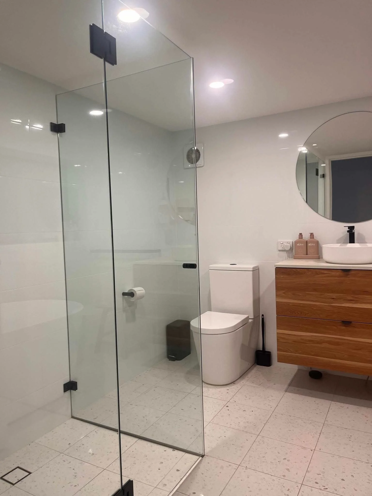 A modern bathroom with a glass shower, a toilet, a wooden vanity with a round sink, a round mirror, two soap bottles, a broom, and a vent on the wall. The bathroom has white walls and tiled floor, with ceiling recessed lights.