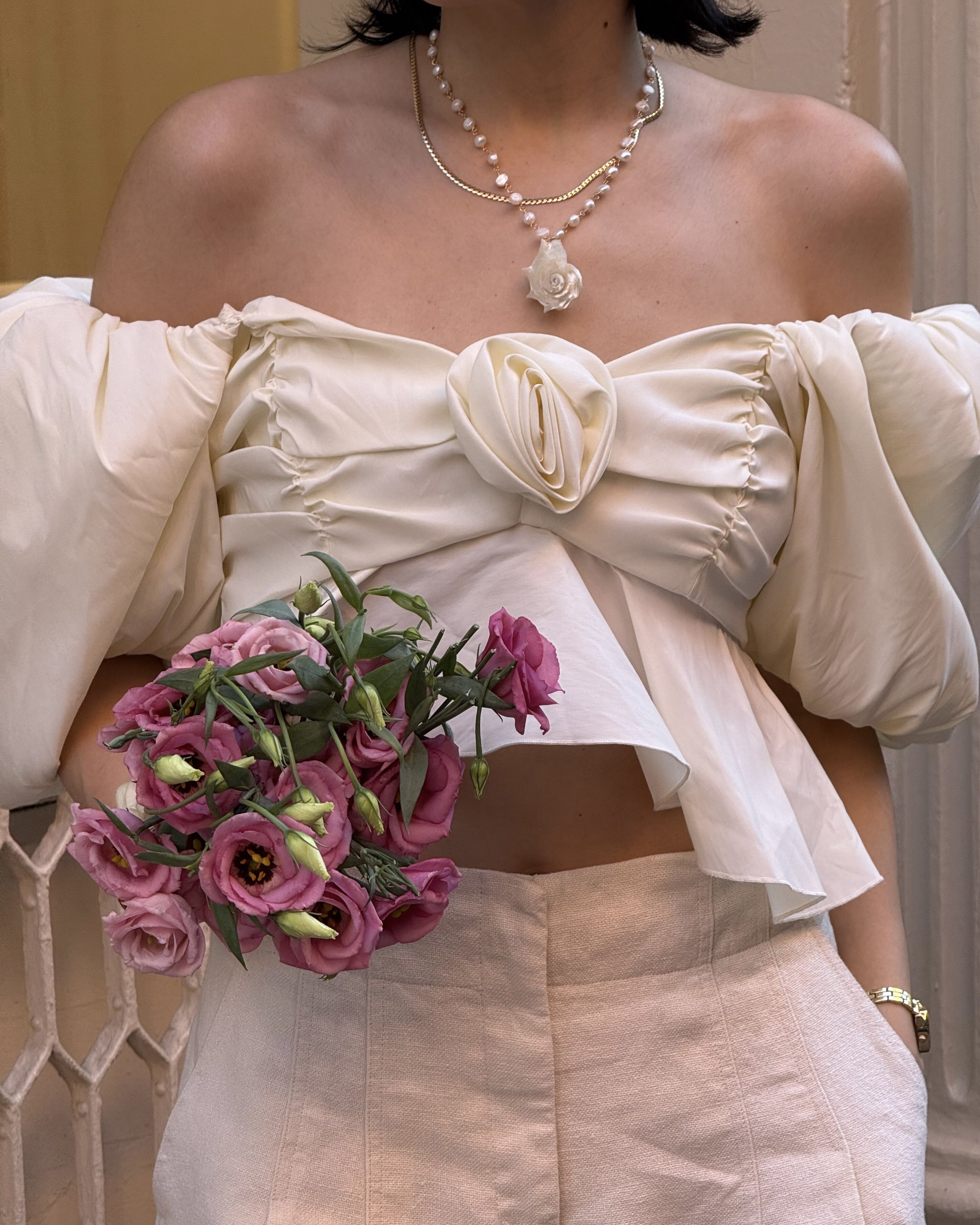 Adriana Alessandra wearing an off-the-shoulder cream blouse with puff sleeves while holding a bouquet of pink flowers in SoHo, New York City.