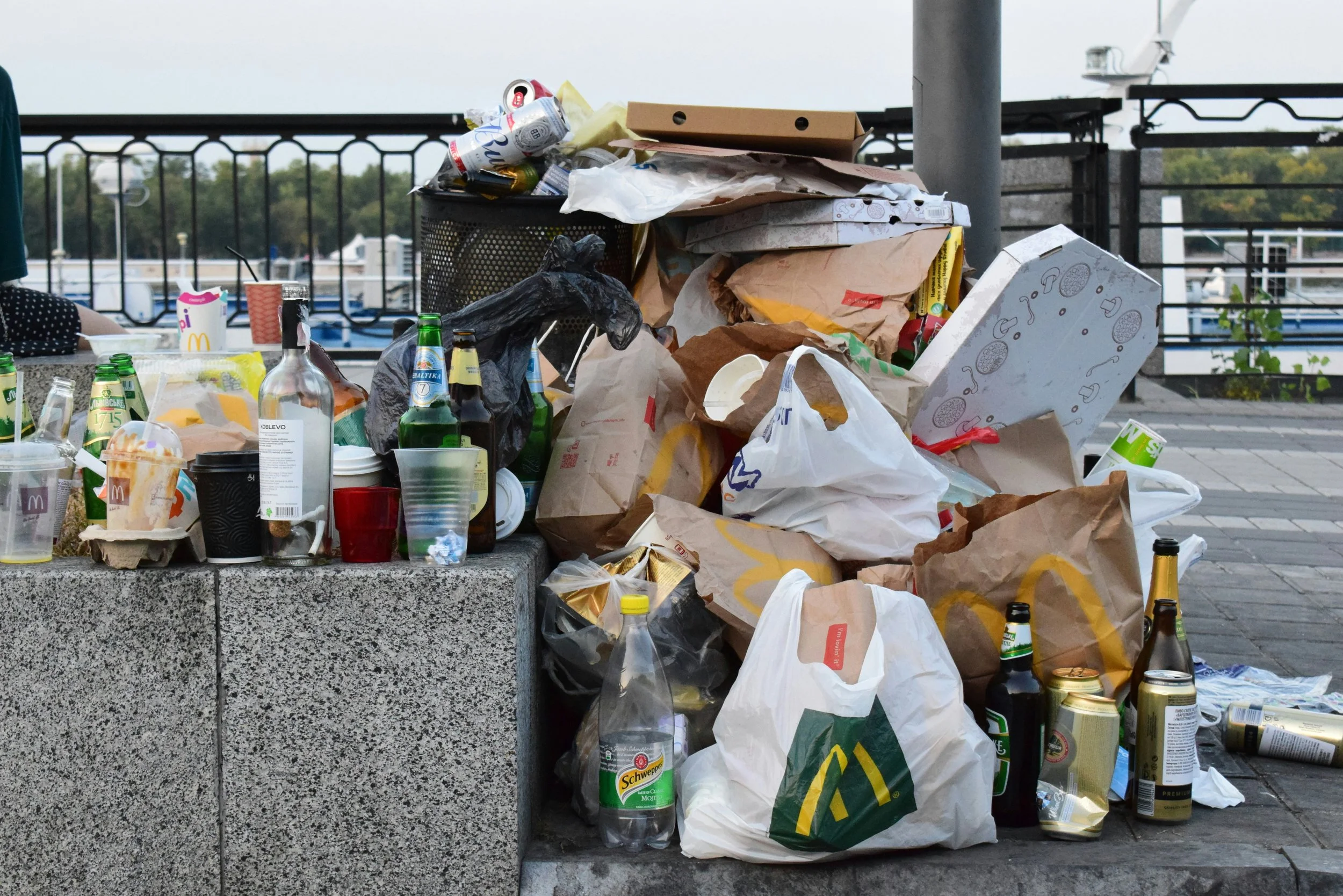 A large pile of garbage and recycling bags and cans on a concrete surface outdoors, with a metal railing and trees in the background.