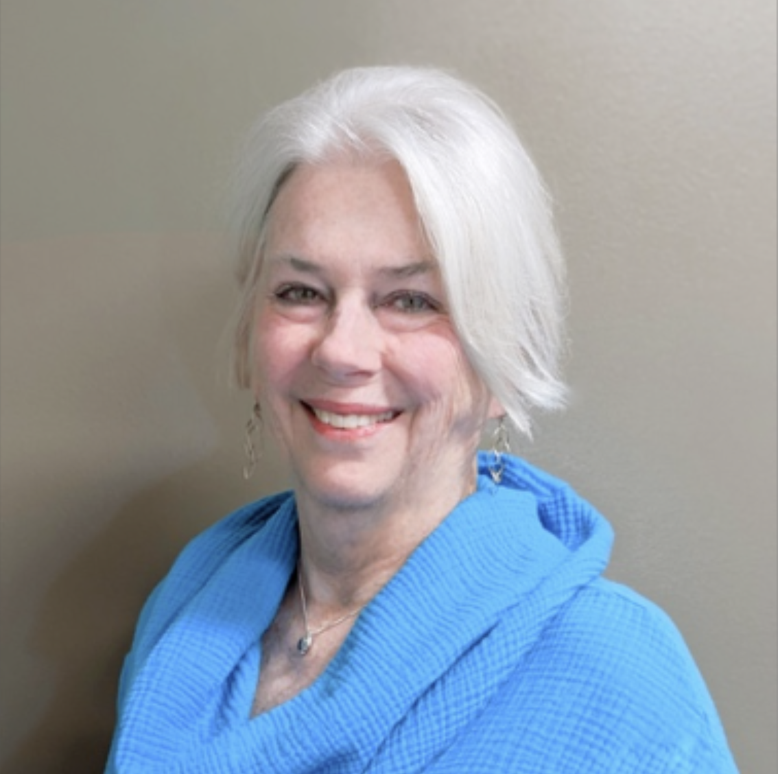 A smiling woman with short white hair, wearing a blue hoodie, and earrings, standing against a plain light-colored wall.