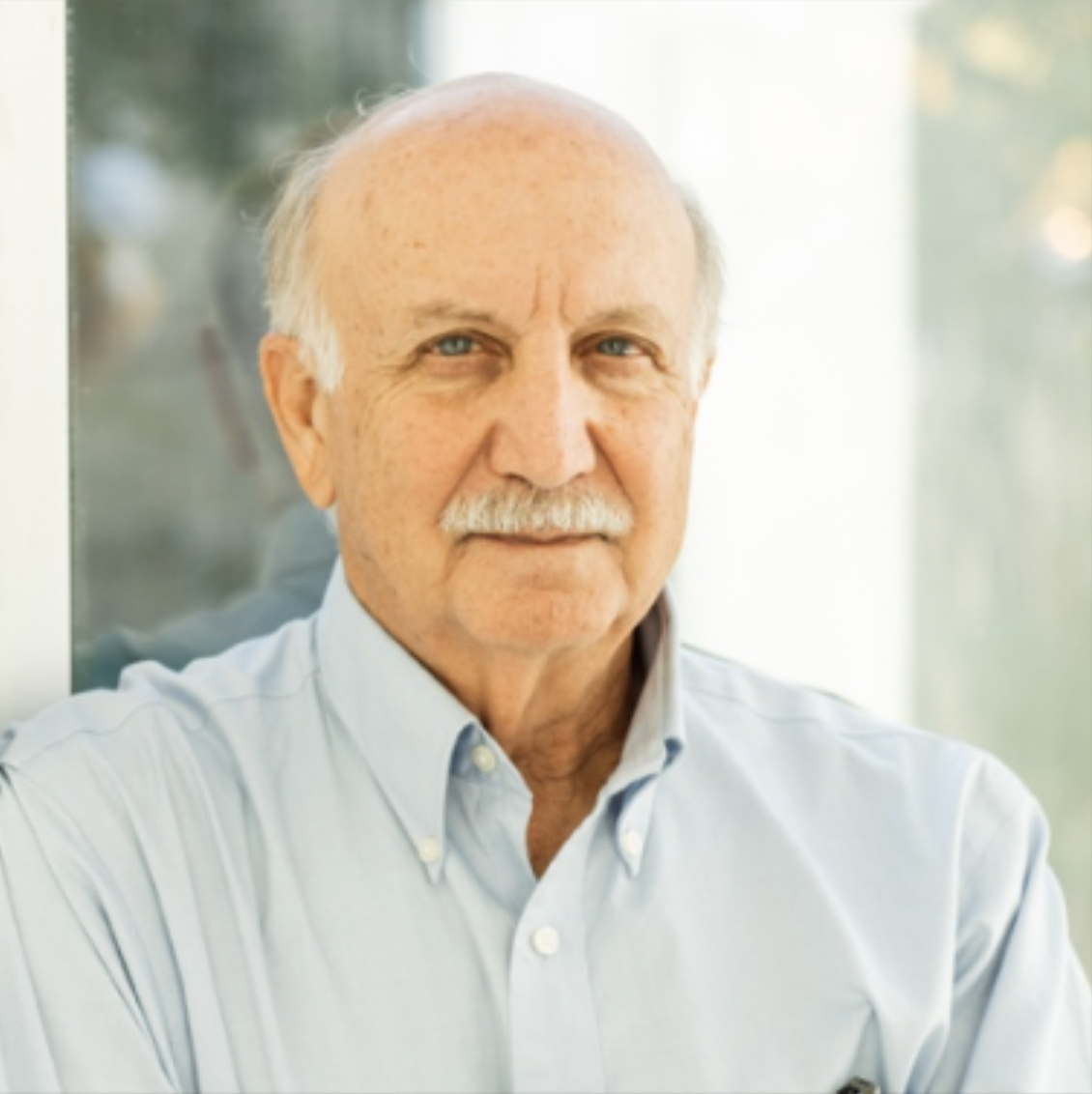 Portrait of an elderly man with a mustache, wearing a light blue button-up shirt, standing indoors with blurred window background.