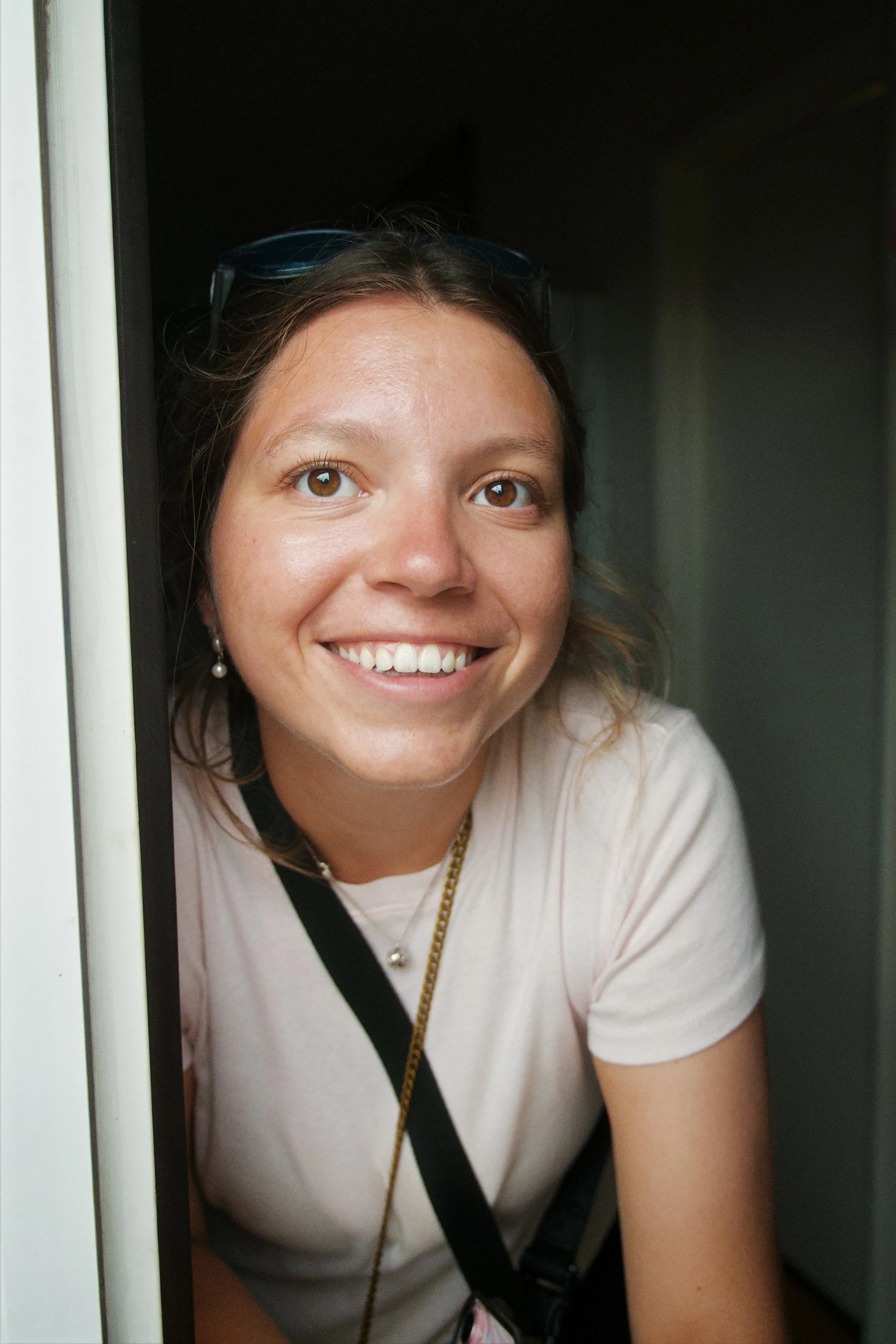 Smiling woman with brown hair, sunglasses on her head, wearing pearl earrings, a pearl necklace, a white t-shirt, and carrying a black strap, peeking through a doorway.