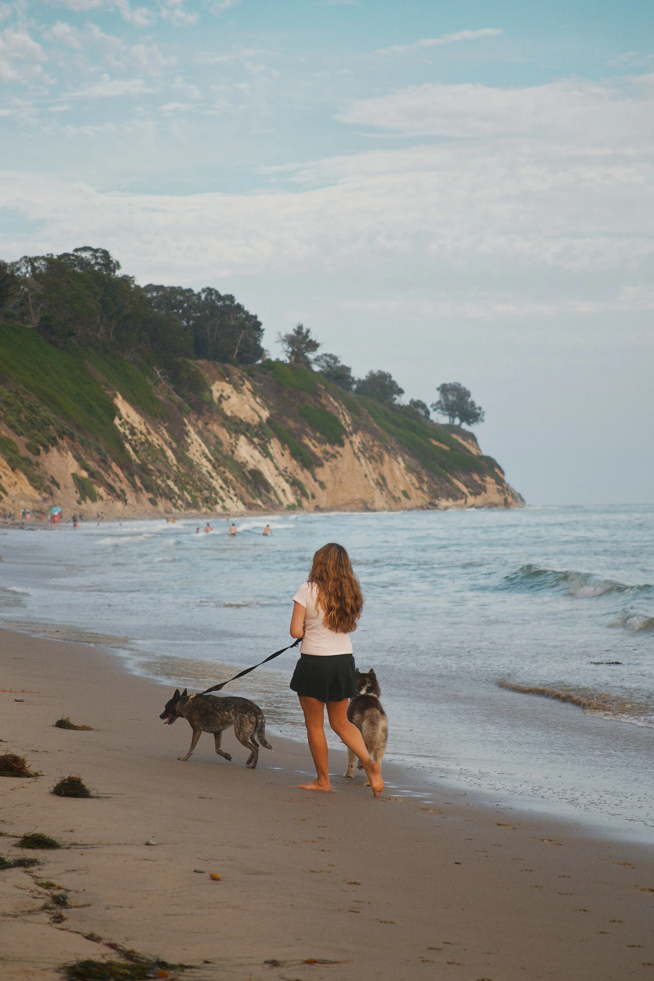 A woman with long red hair, wearing a light-colored shirt and black shorts, walking two dogs on a leash along the sandy shoreline of the ocean with cliffs and trees in the background.