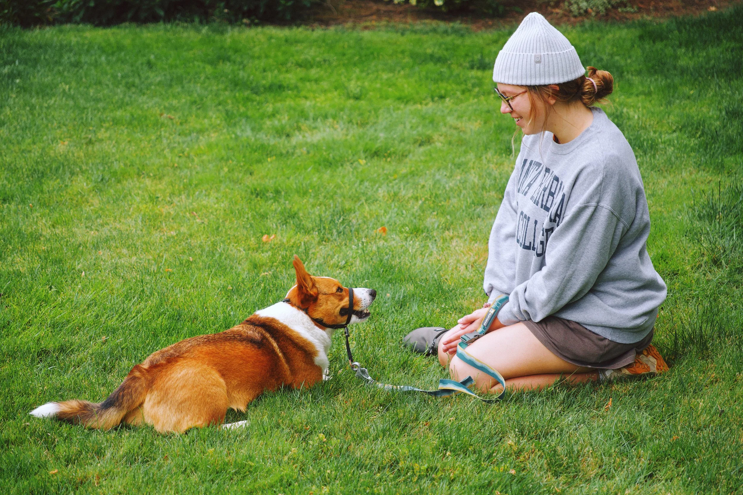 A young woman with red hair, glasses, and a gray beanie, sitting on the grass and smiling at a brown and white dog. The dog is lying down with a leash attached, and they are outdoors on a green lawn.