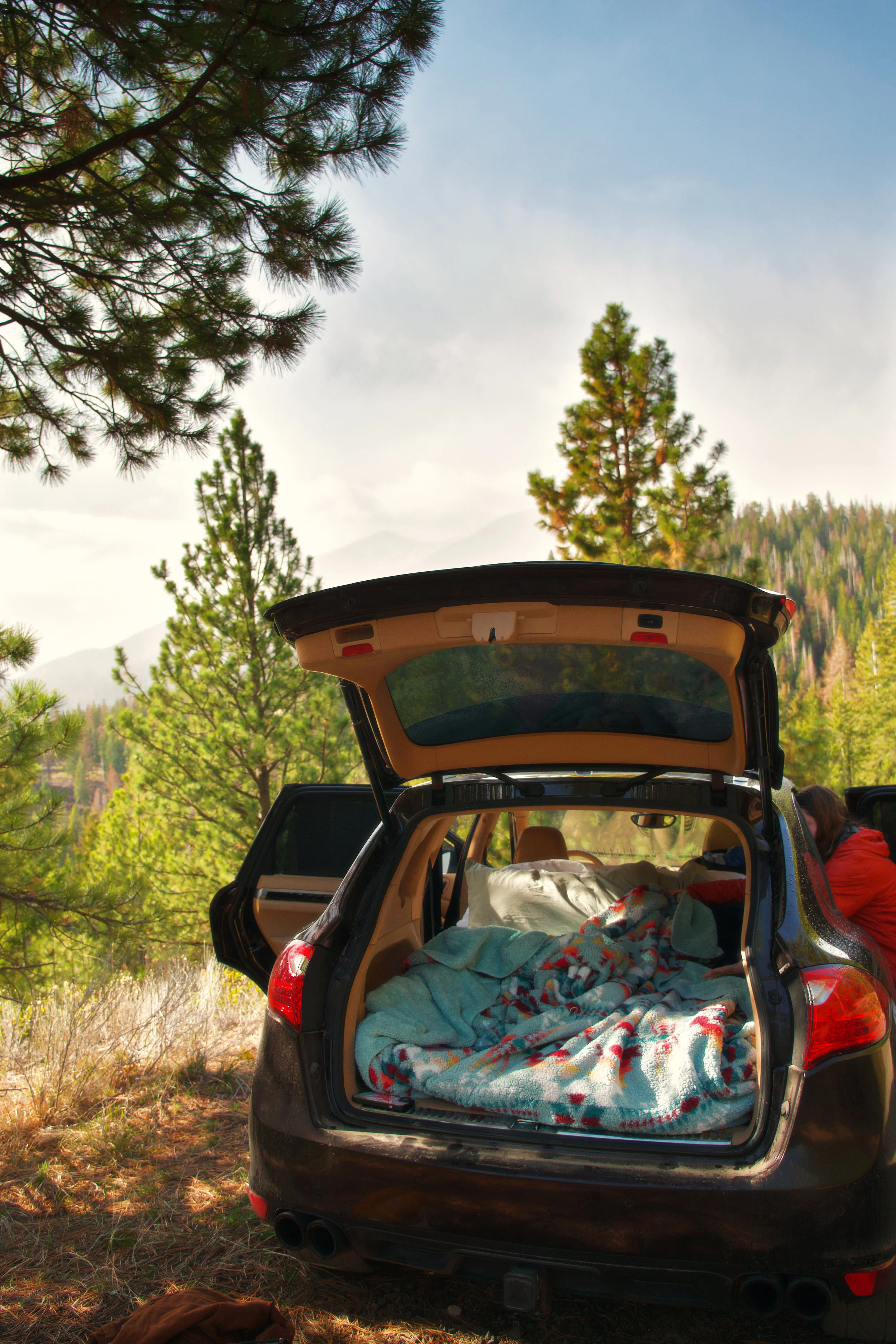 The rear of a black SUV with its trunk open, showing a bed with blankets and pillows inside. Two people sit on the ground beside the vehicle in a wooded outdoor setting with tall trees and mountains in the background.