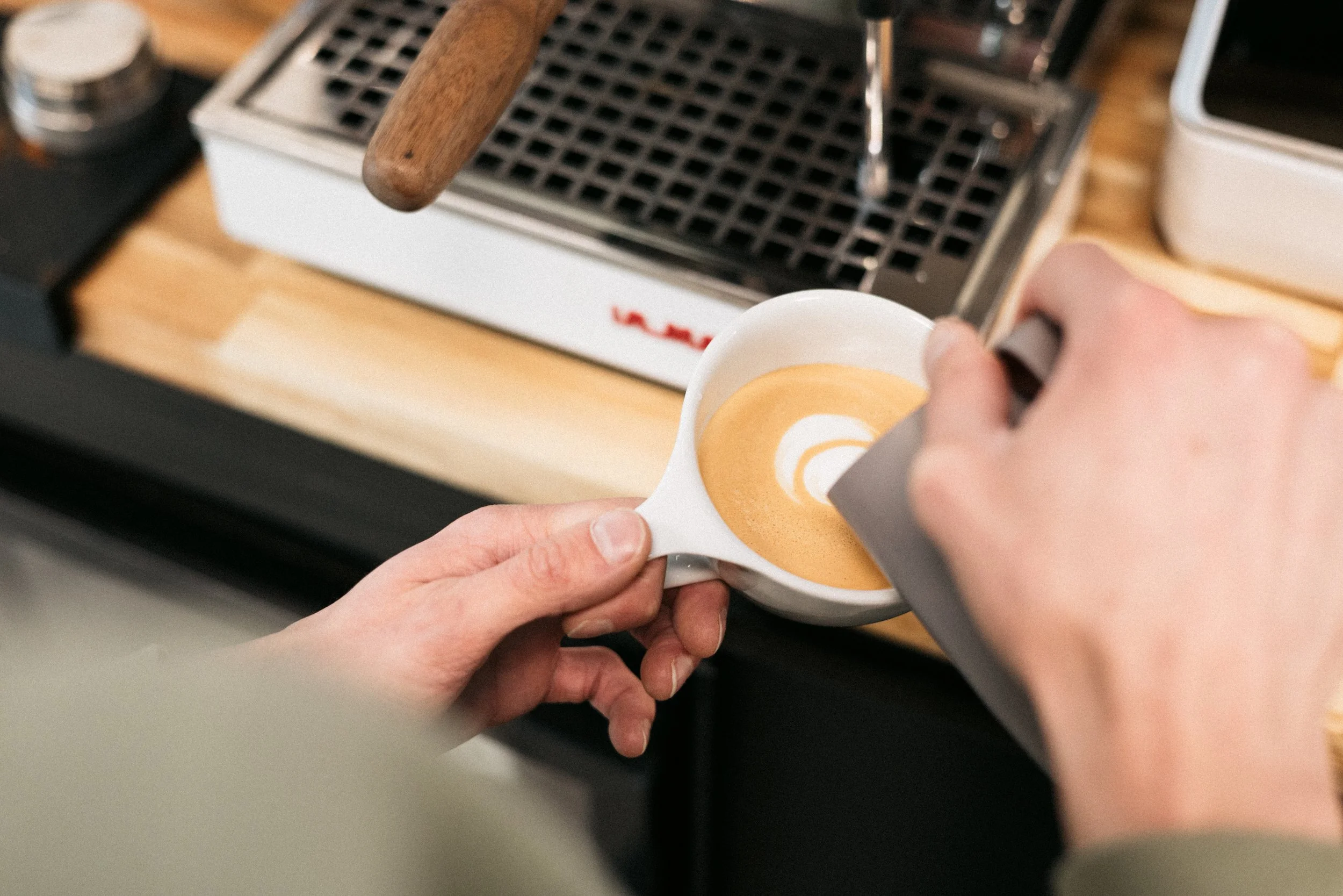 Person pouring coffee into a cup on a kitchen countertop with a coffee machine in the background.