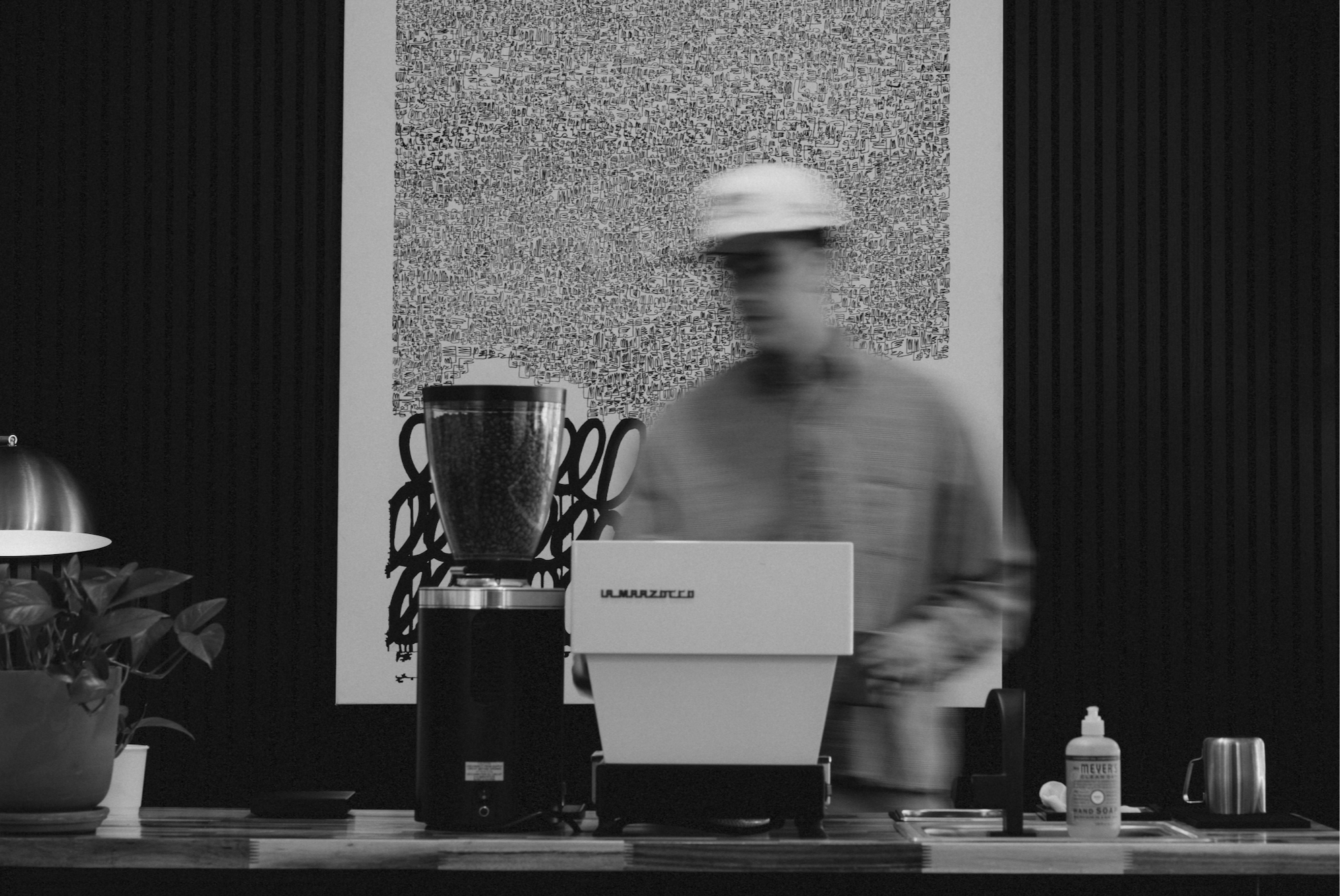 A person preparing coffee using an espresso machine at a counter. The background features an abstract patterned wall, and there is a potted plant on the left side of the counter. The photo appears blurry with motion, likely taken in black and white.