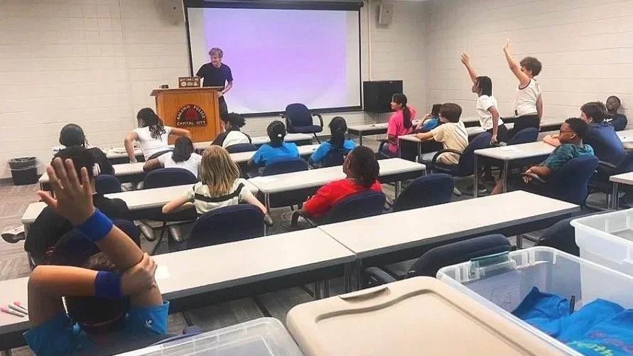 A classroom with children sitting at desks, some raising their hands, facing Andy at a podium and a whiteboard.