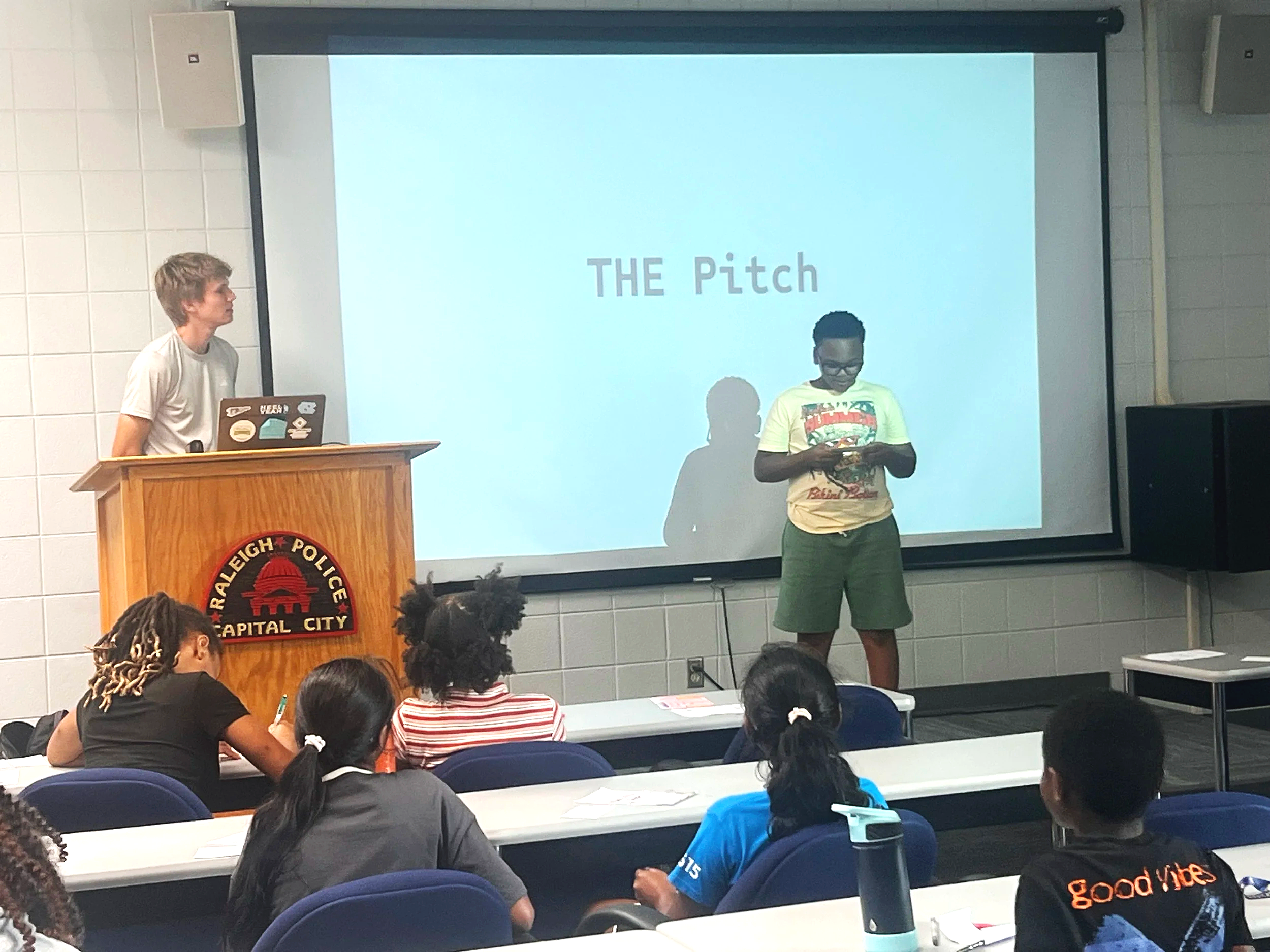 A classroom presentation shows boys standing at the front, one behind a wooden podium with a laptop, and the other to the right, holding a phone. A large screen behind them displays "THE Pitch". Students are seated at desks, facing the front, with some taking notes.