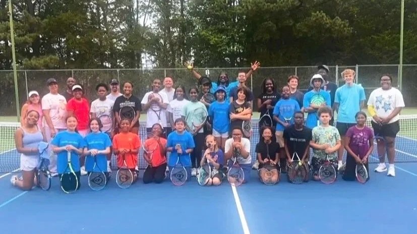 Group of children and teenagers with coaches on a tennis court, holding tennis rackets, during daytime.