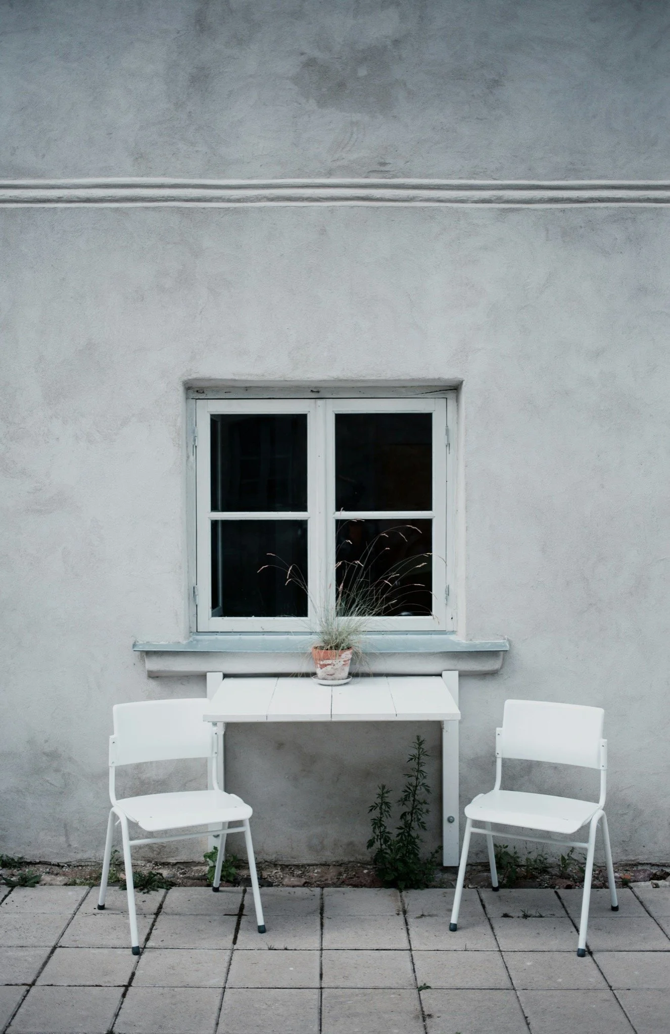 Outdoor patio with white table, two white chairs, potted plant on table, window on light gray wall, and tiled ground with some weeds.