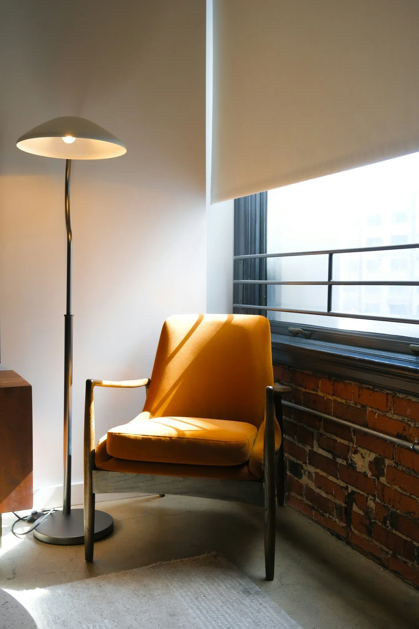 A cozy corner with a mustard yellow armchair, a modern black floor lamp, a window with partially rolled-up blinds, and a brick wall with a metal railing outside.