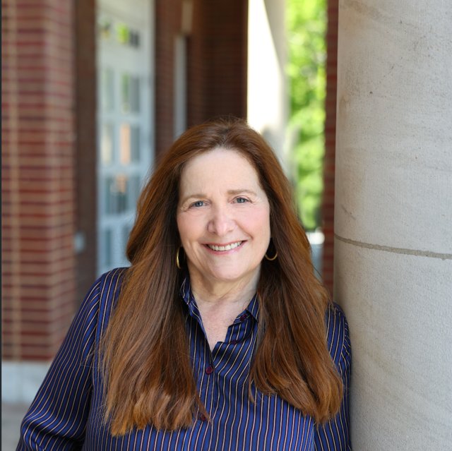 A woman with long red hair smiling, wearing a blue striped shirt, standing outdoors next to a large column with a brick building and green trees in the background.
