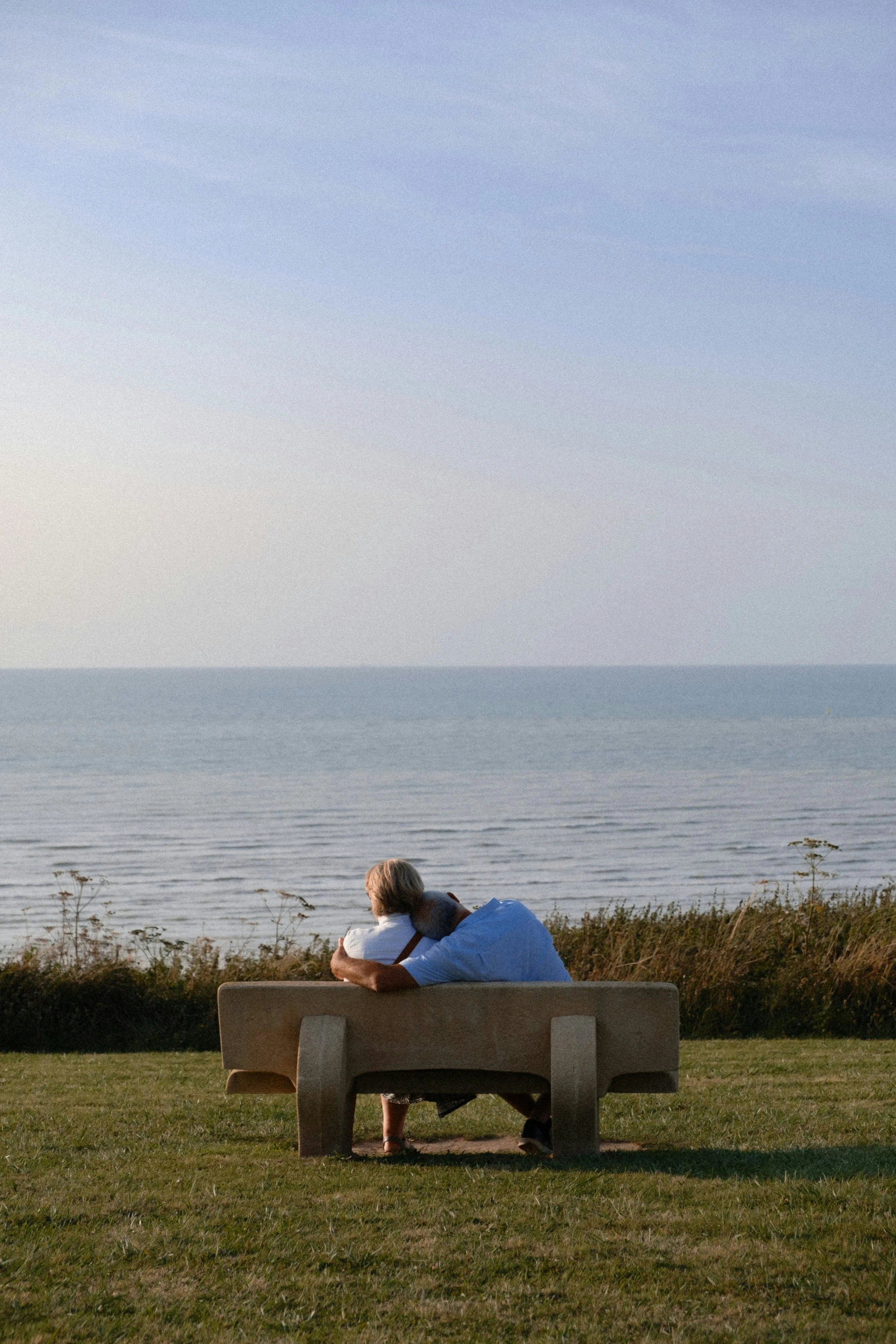 Two people sitting on a bench near a body of water, embracing as they look at the horizon with a cloudy sky above during sunset.