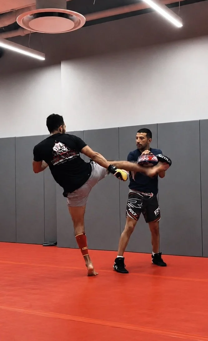 Two men practicing martial arts in a gym, one executing a kick while the other holds focus pads. The gym has gray walls, red floor, and modern ceiling lights.
