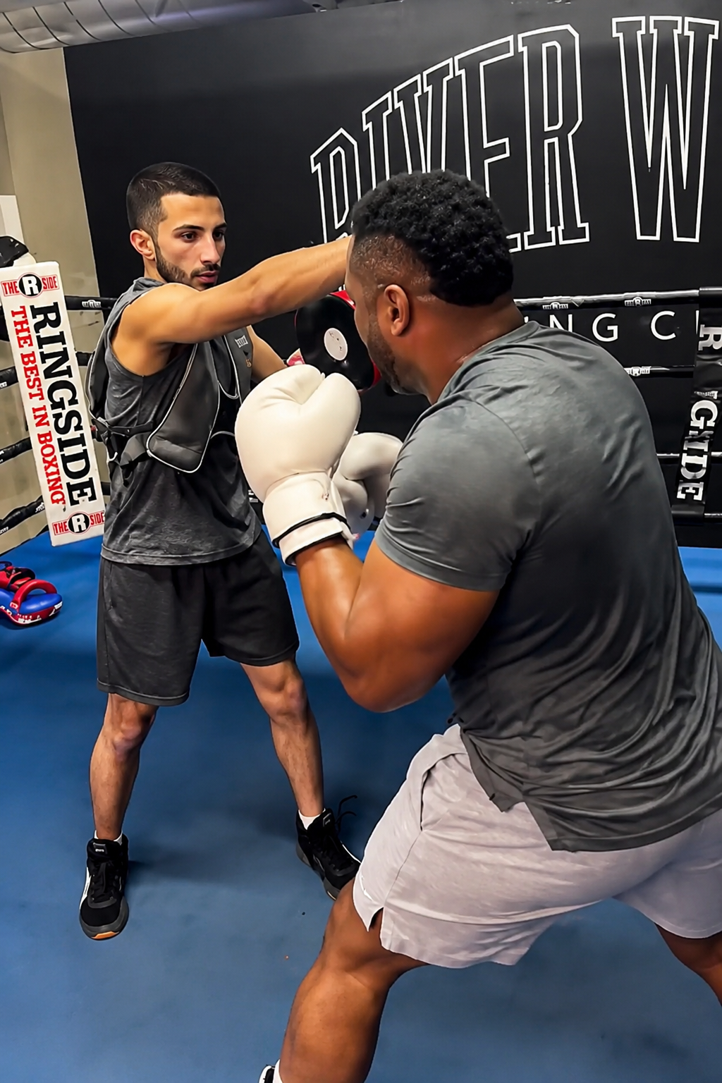 Two men training in a boxing gym, one is throwing a punch while the other is in a defensive stance, wearing boxing gloves.