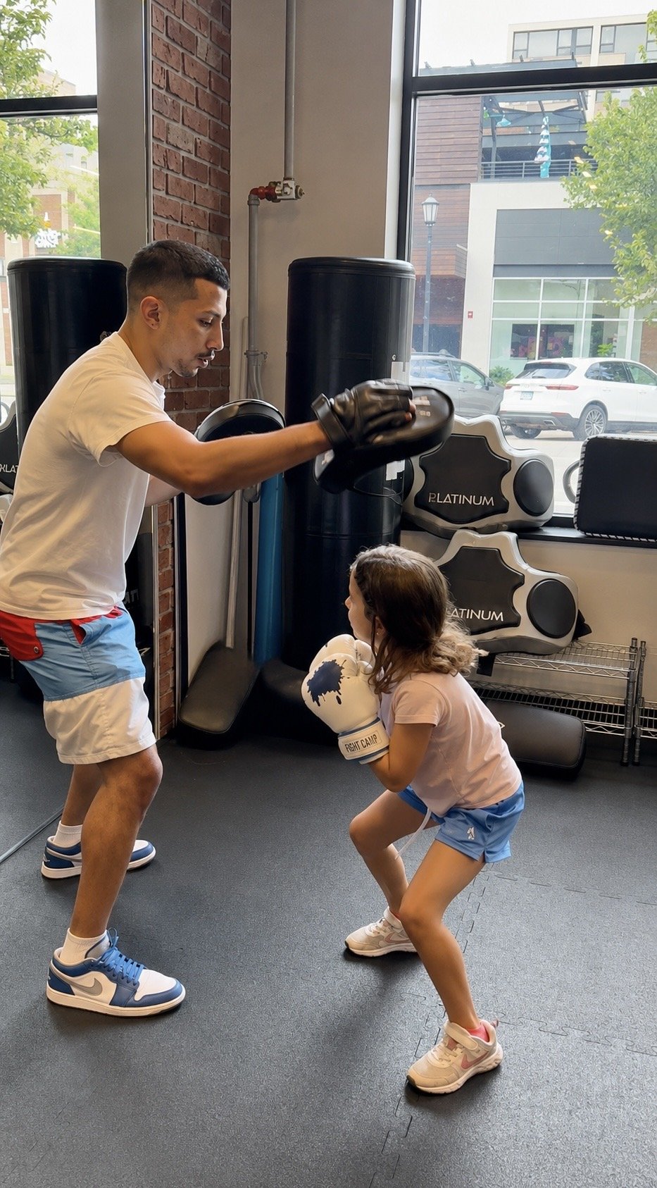 A young girl wearing boxing gloves is practicing boxing with a trainer in a gym. The trainer is holding focus pads in front of him while the girl is in a boxing stance. The gym has large windows, boxing equipment, and a brick wall.