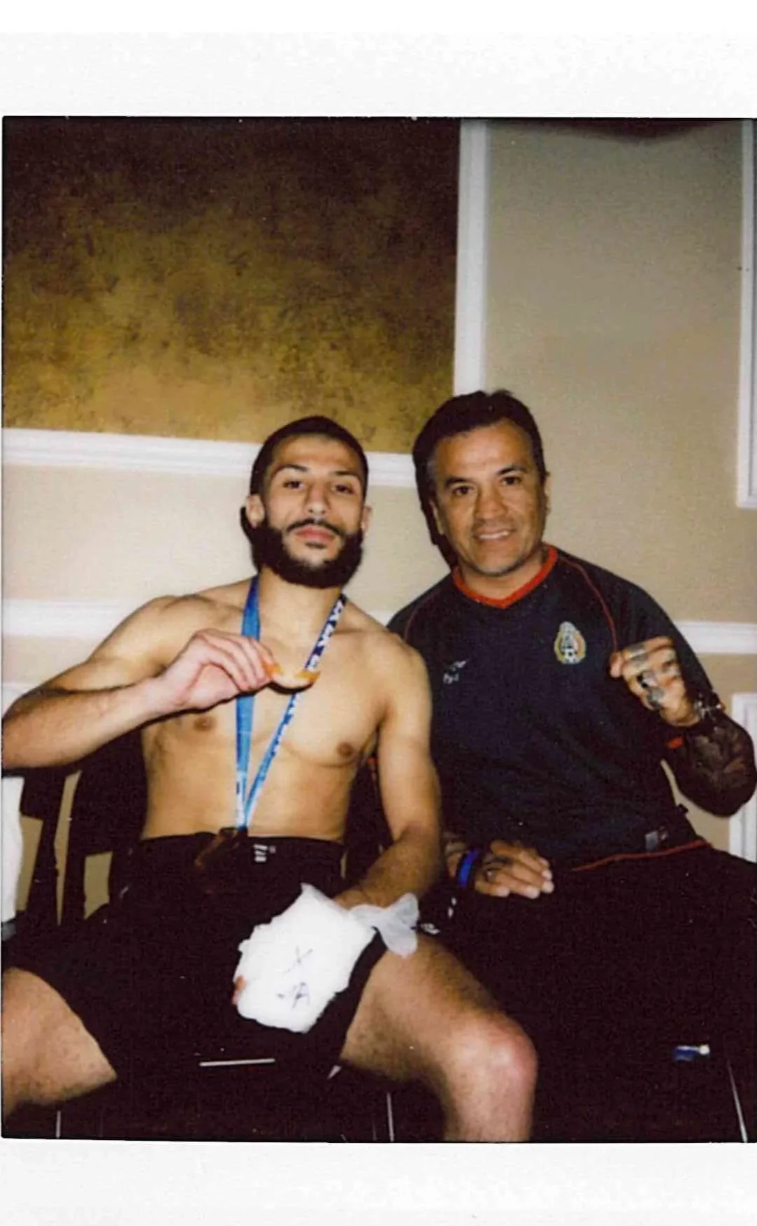 Two men, one shirtless and wearing a medal, and the other in a sports jersey, sitting together in a room with a gold and beige wall background.