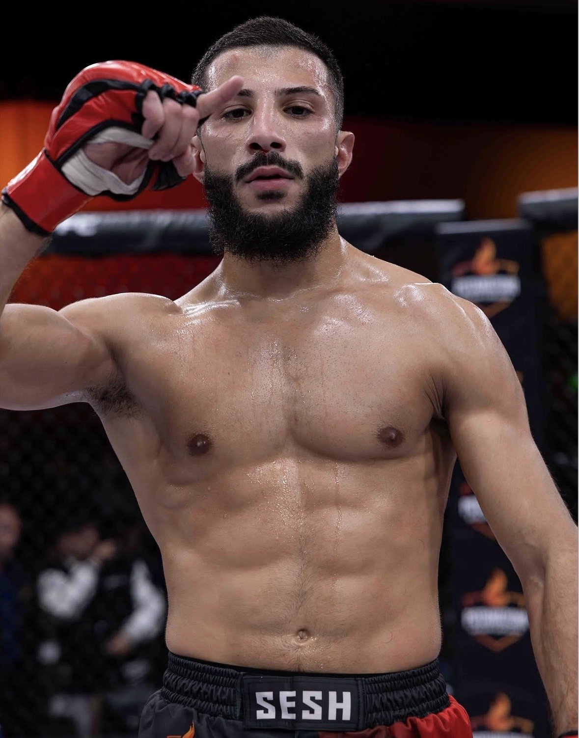 A shirtless man with a beard and sweat on his face, inside a mixed martial arts cage, wearing red and black fight shorts with the word 'SESH' on the waistband, holding his right hand near his face in a fist.