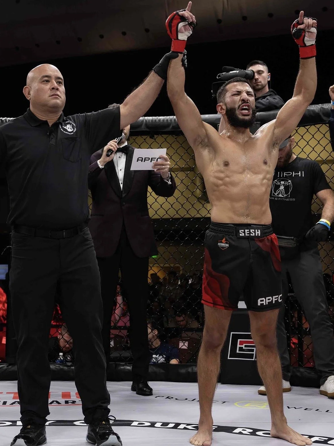 Mixed martial arts fighter in the cage celebrating victory with raised arms, referee holding his hand, others in background, octagon cage.