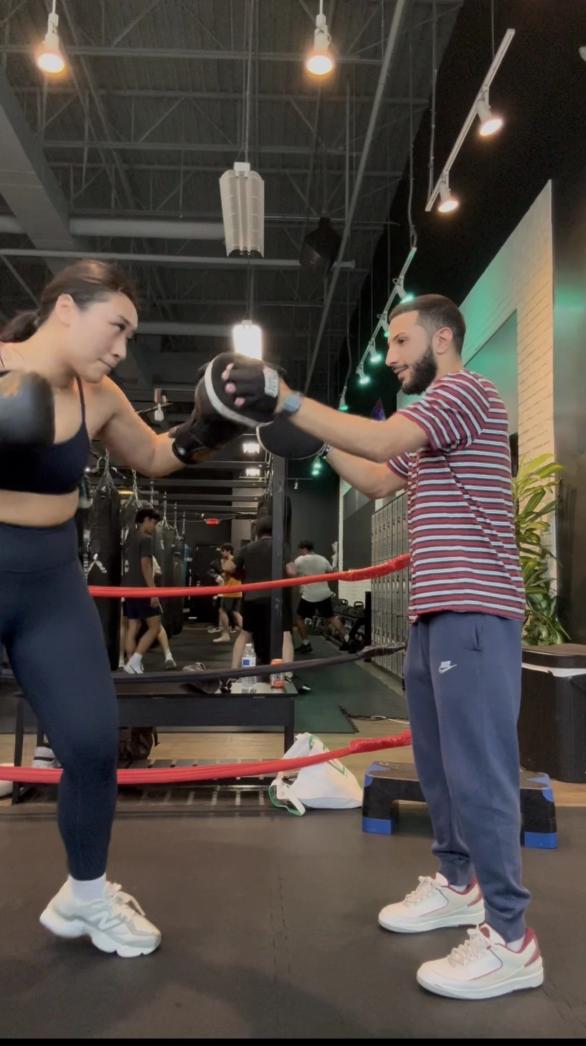A man and a woman practicing boxing in a gym. The woman is wearing boxing gloves, and the man is holding and guiding her punches. They are standing inside a boxing ring with other people exercising in the background.