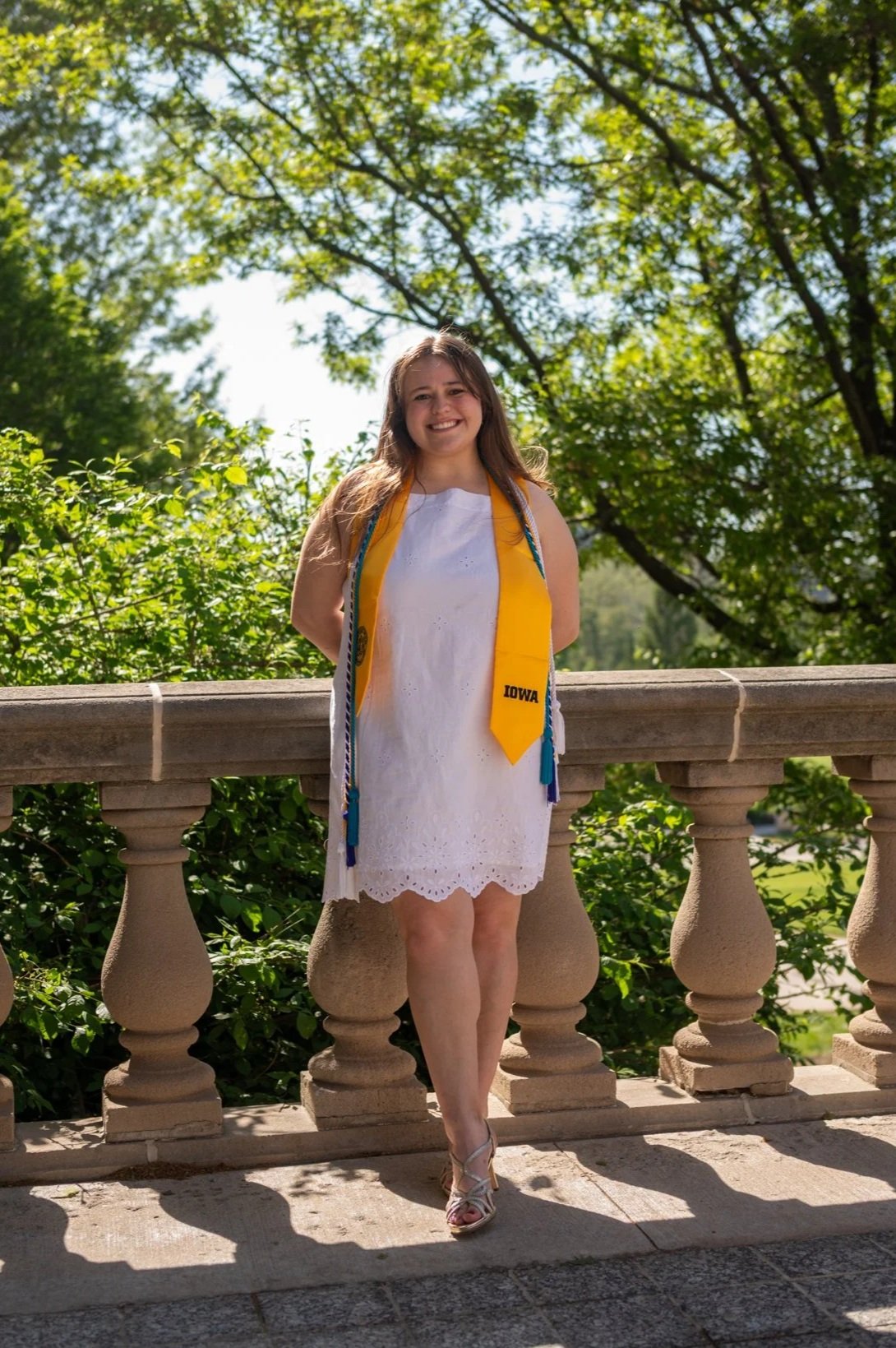 A young woman wearing a white dress and a yellow sash that says 'IOWA' stands outdoors on a stone balcony with decorative balusters. She is smiling, with her hands behind her back, and there is lush green foliage and trees in the background. The photo was taken on a sunny day.