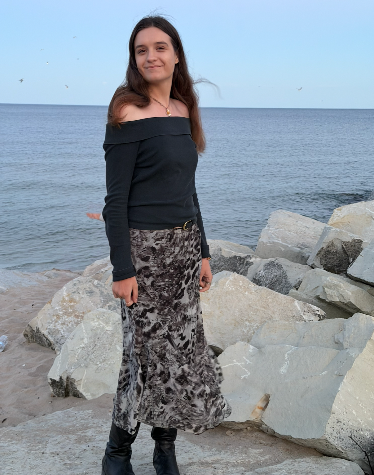 A woman standing on rocks by the ocean, wearing a black off-the-shoulder top and a long animal print skirt.