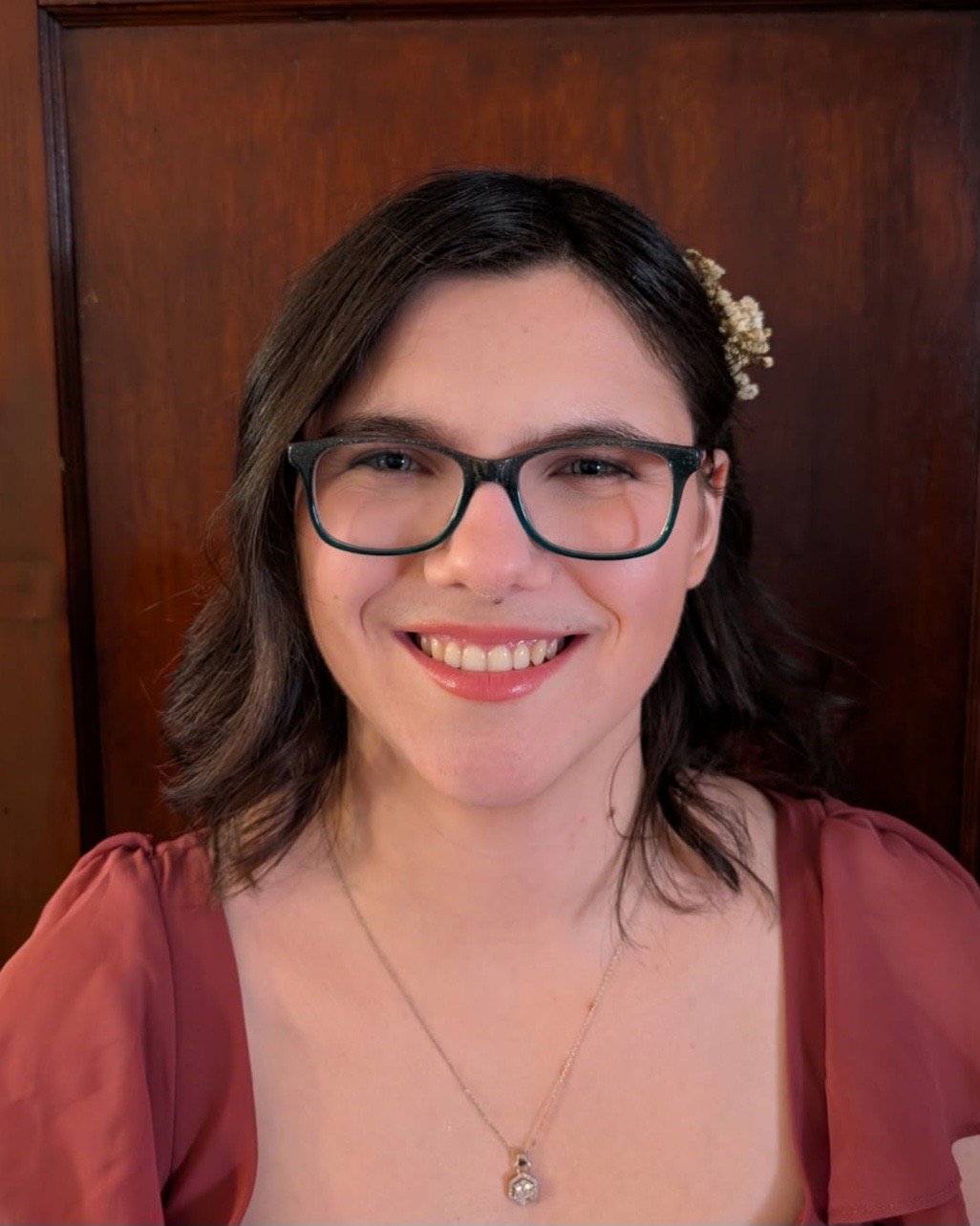A woman with glasses and dark, wavy hair smiling in front of a wooden background, wearing a reddish-brown dress and a necklace with a pendant, and a small white flower in her hair.