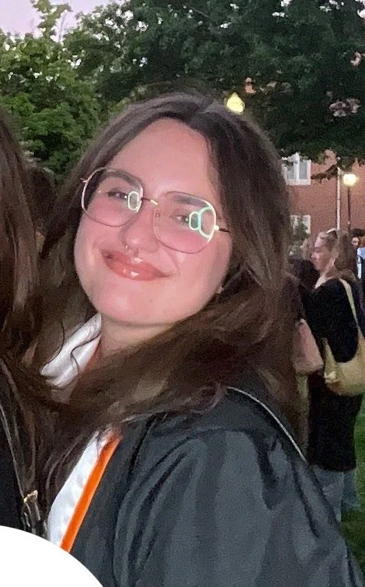A young woman with glasses and long brown hair smiling outdoors during twilight, with trees and people in the background.