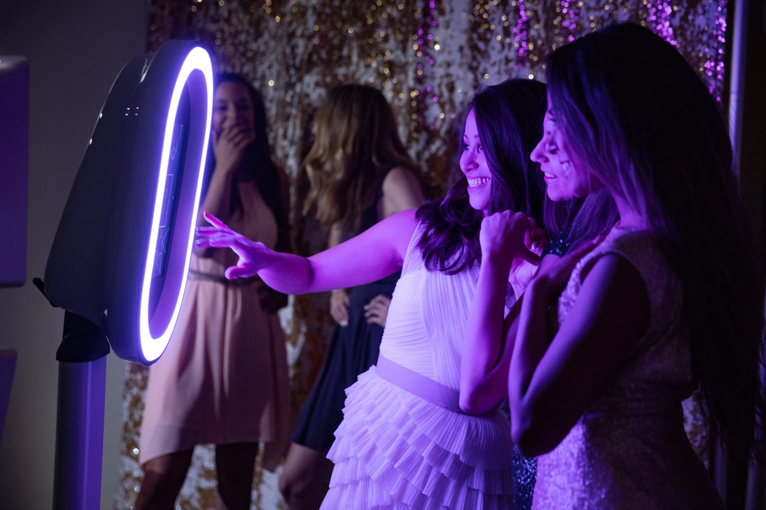 Women at a party using a photo booth with purple neon lighting, smiling, and enjoying themselves.