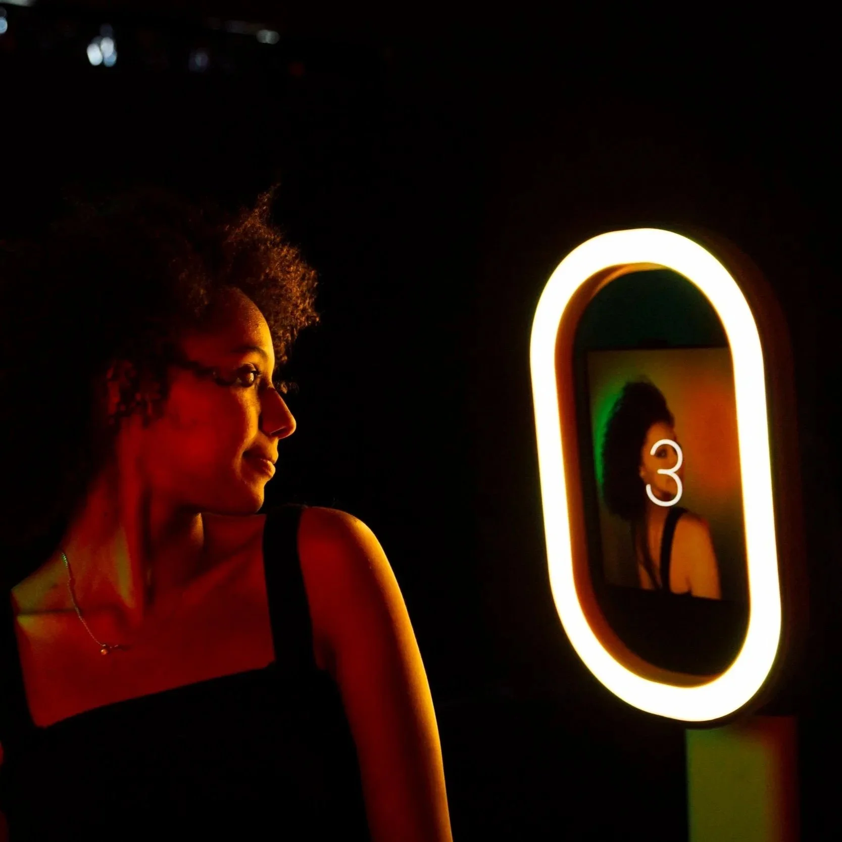A woman with curly hair looking at her reflection in a mirror with a neon light frame, in a dimly lit room.