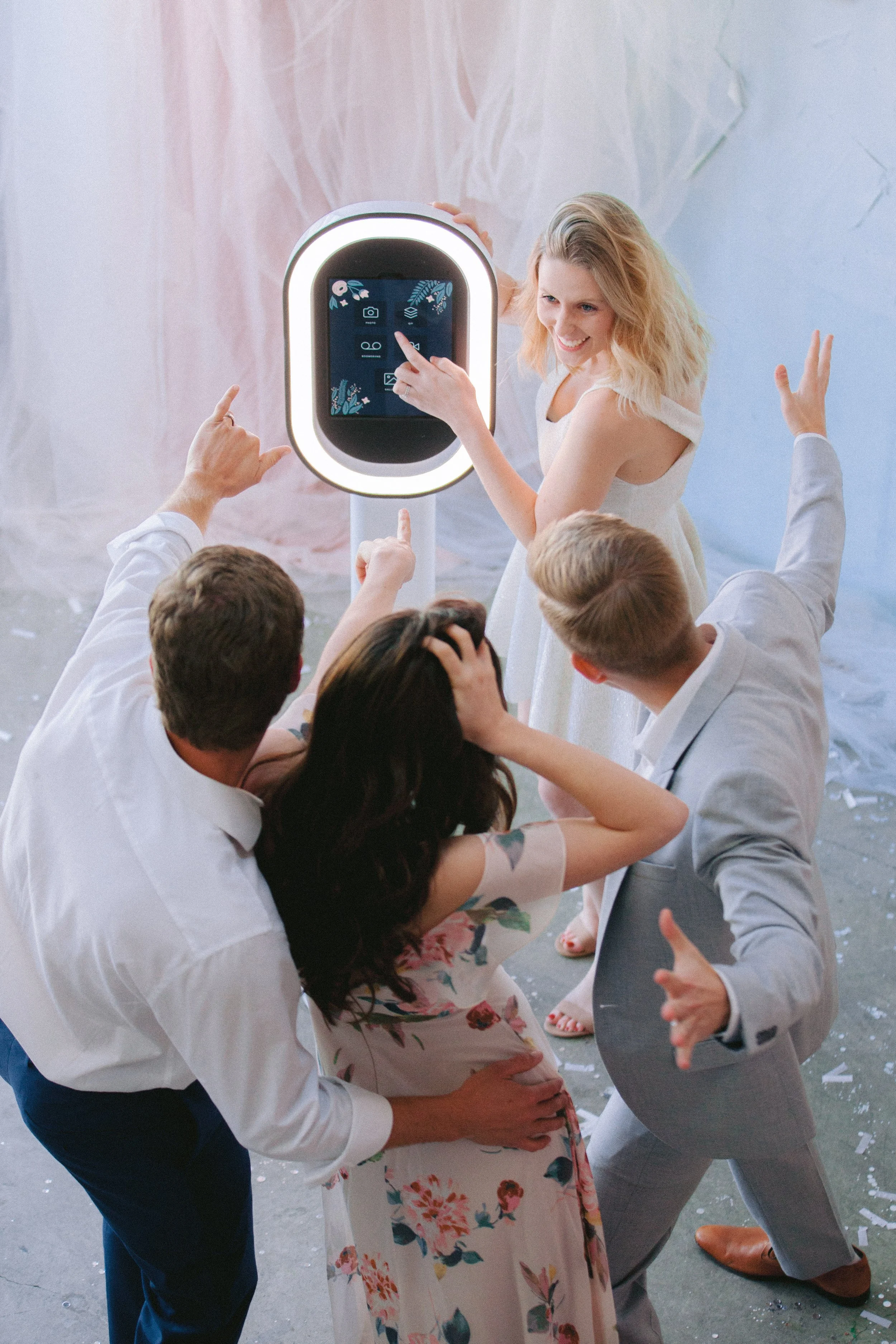 A group of people dancing at wedding reception, with some pointing and others hugging, while a woman in a floral dress and a man in a gray suit smile and engage with each other.