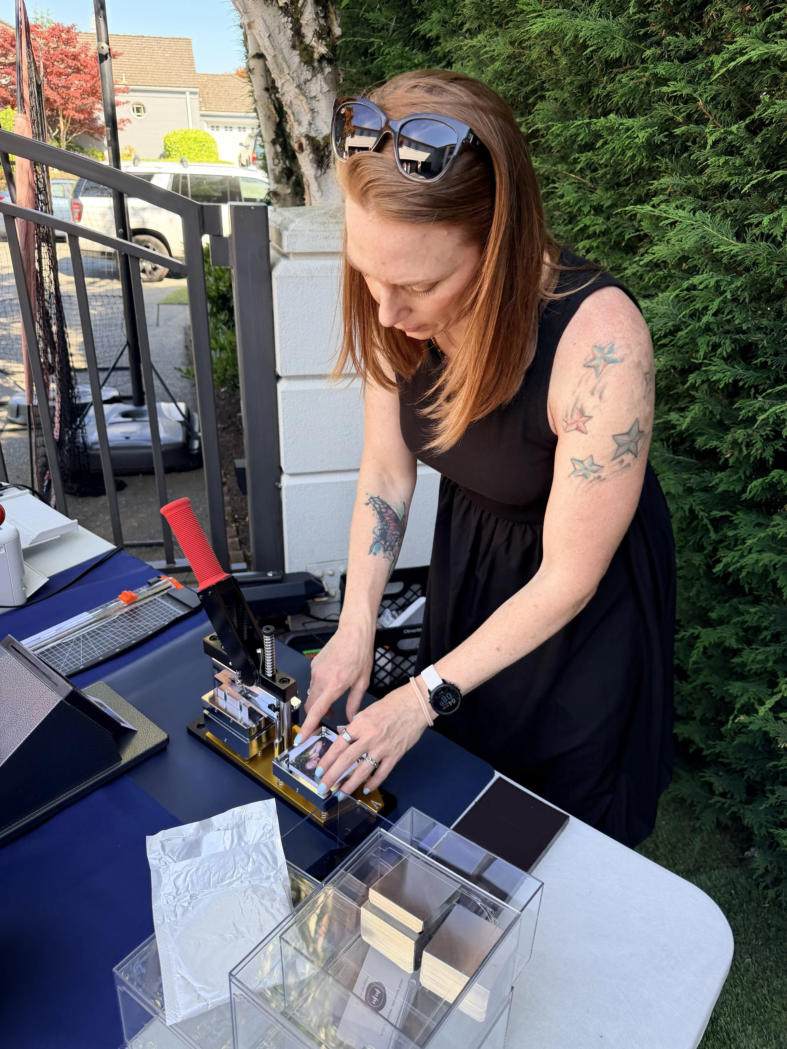 A woman with red hair, tattoos on her arms, wearing sunglasses on her head, working on a small model or device at an outdoor table with various boxes and instruments, green hedge and trees in the background.