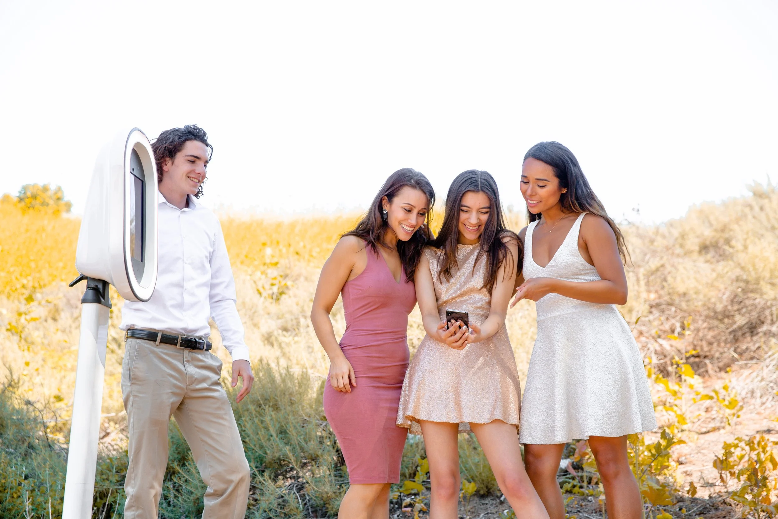 Four young women and one young man looking at a smartphone outdoors in a field with yellow flowers, with a piece of scientific equipment nearby.