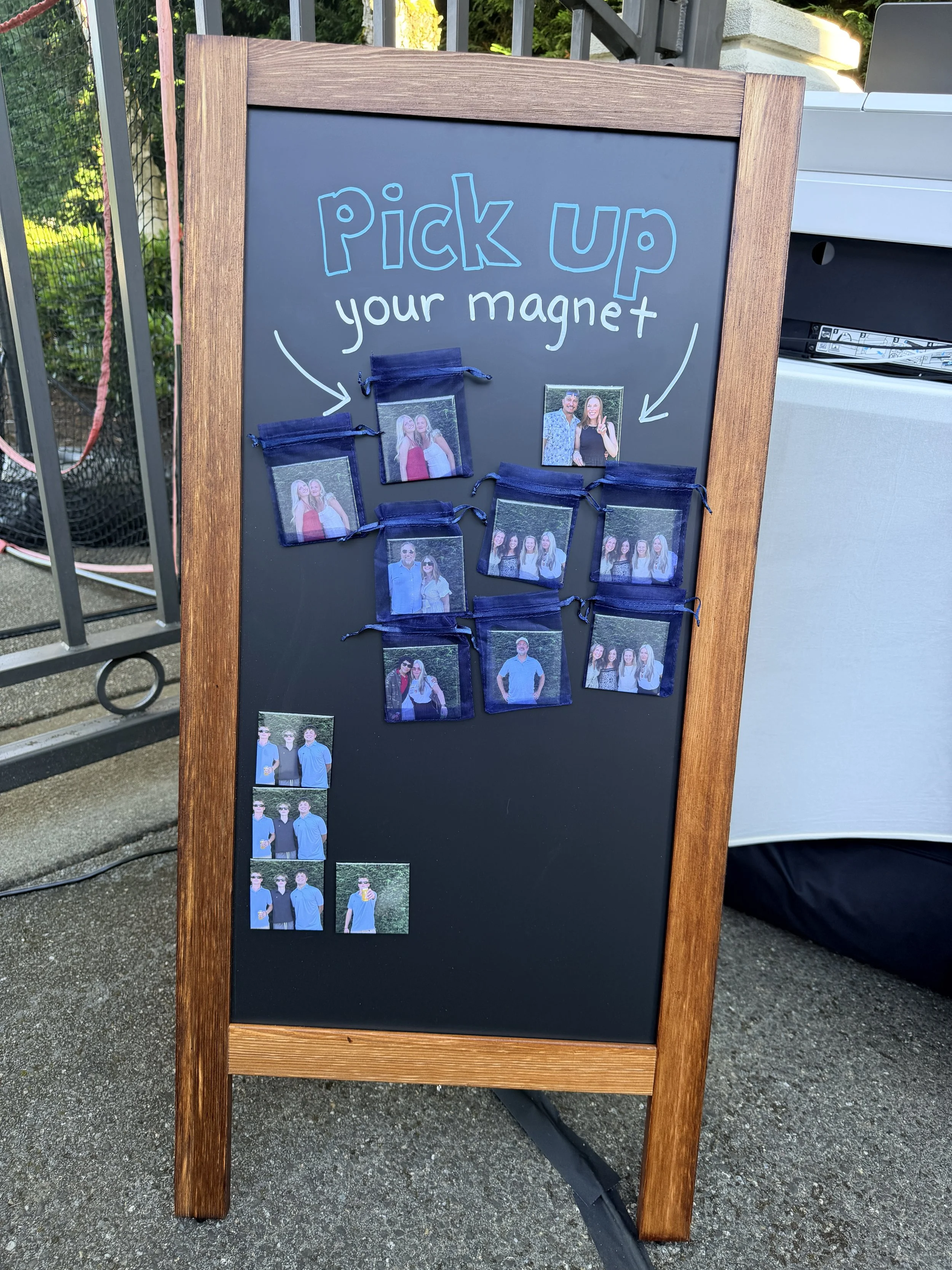 A black chalkboard sign with a wooden frame, displaying small photos of people. The sign reads 'Pick up your magnet' with handwritten arrows pointing to some of the photos.
