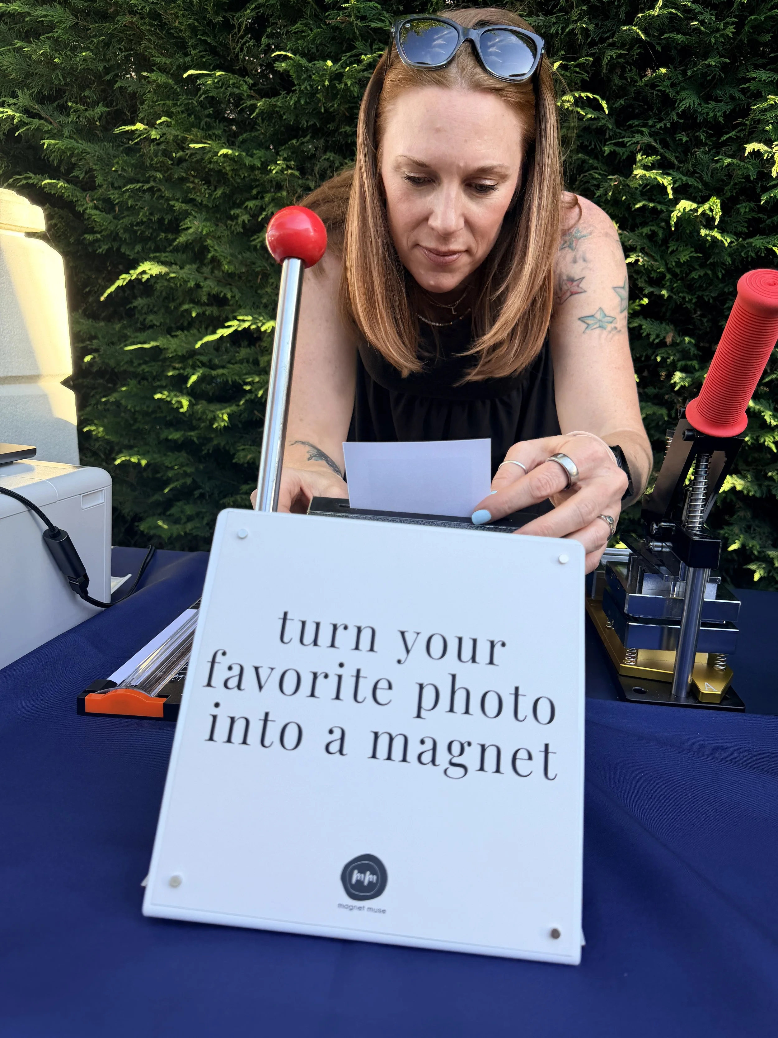 A woman with red hair and tattoos on her arms sitting outdoors at a table with photo transfer equipment, including a magnetic photo transfer device, a red roller, and other tools, preparing to turn a photo into a magnet.