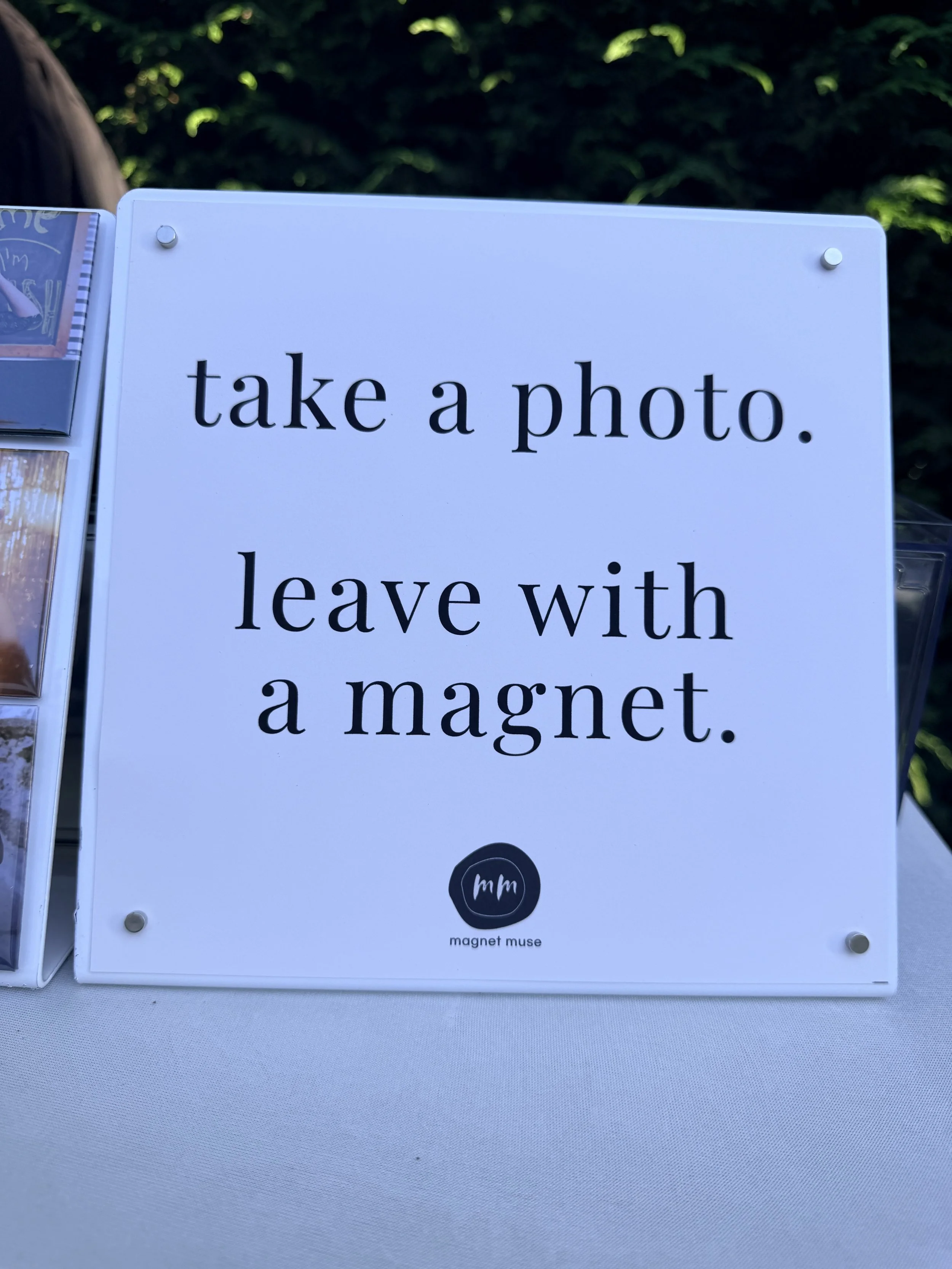 A white sign with black text that reads 'take a photo. leave with a magnet.' At the bottom, there is a small round logo with letters 'mm' and the words 'magnet muse' underneath.