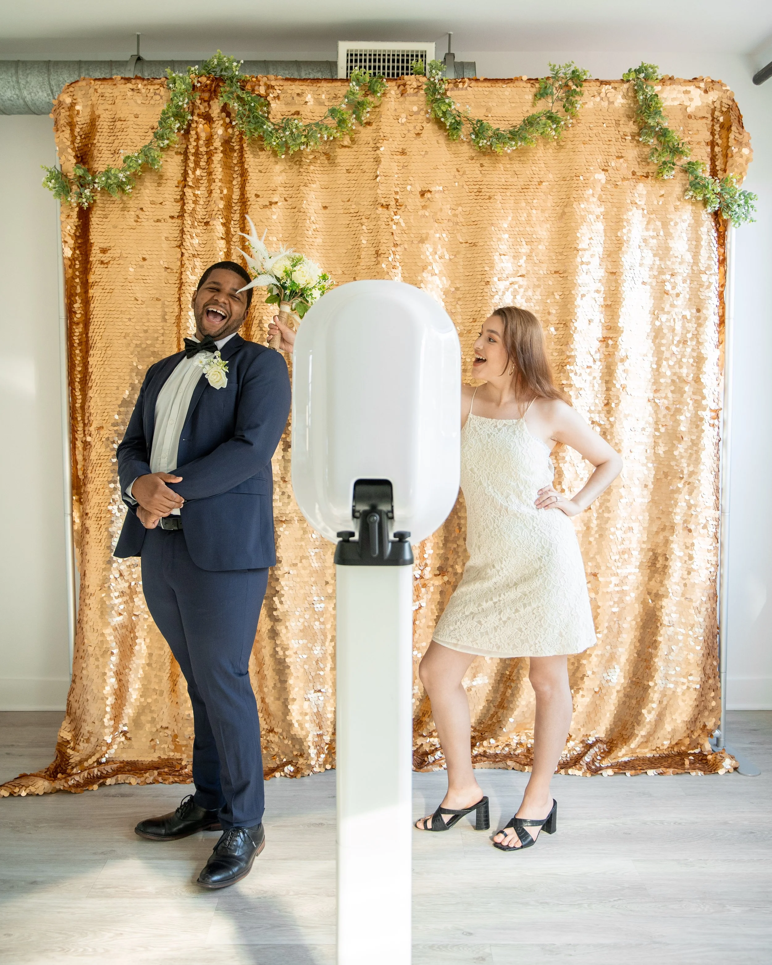 A happy man in a wedding tuxedo and a woman in a white dress posing in front of a gold sequin backdrop for photos, with a photo booth camera in front of them.