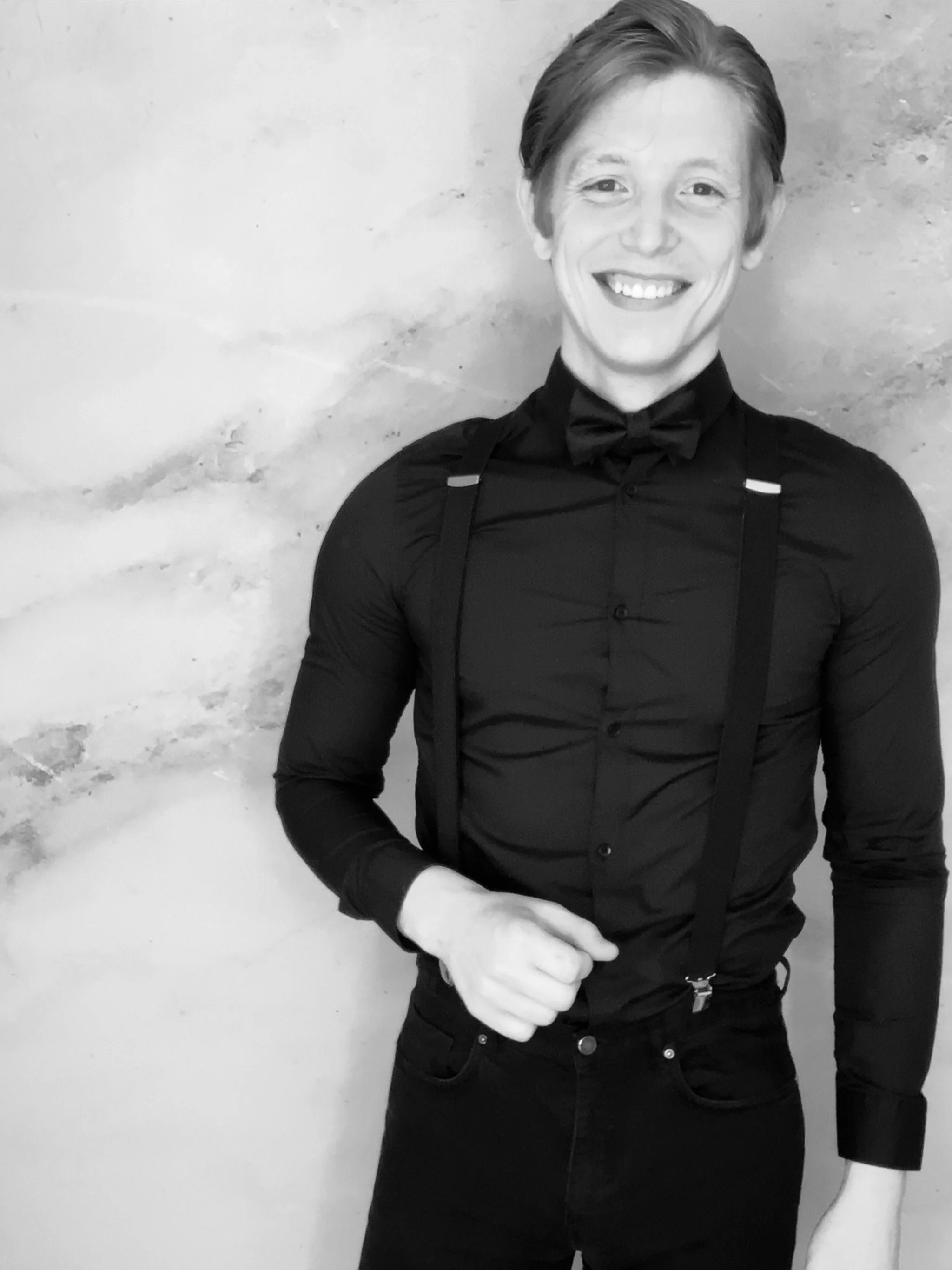 A young man smiling, wearing a black shirt, black bow tie, and suspenders, standing against a plain wall.