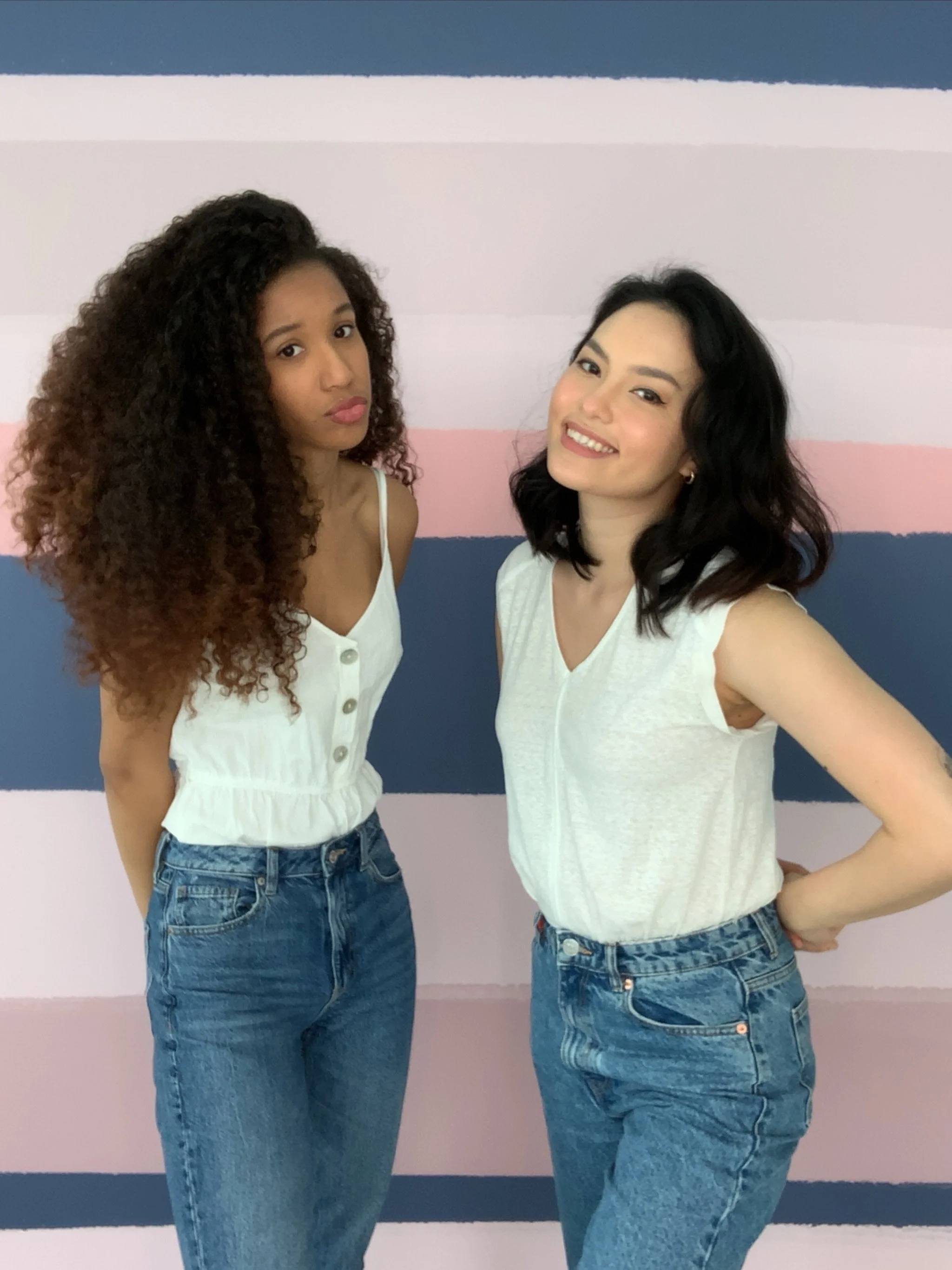 Two women standing in front of a striped pastel pink, blue, and white wall. The woman on the left has curly brown hair and is wearing a white sleeveless top with buttons and high-waisted blue jeans. The woman on the right has black wavy hair and is wearing a white sleeveless top and high-waisted blue jeans. They are posing casually, with the woman on the right smiling and the woman on the left making a kissing face.