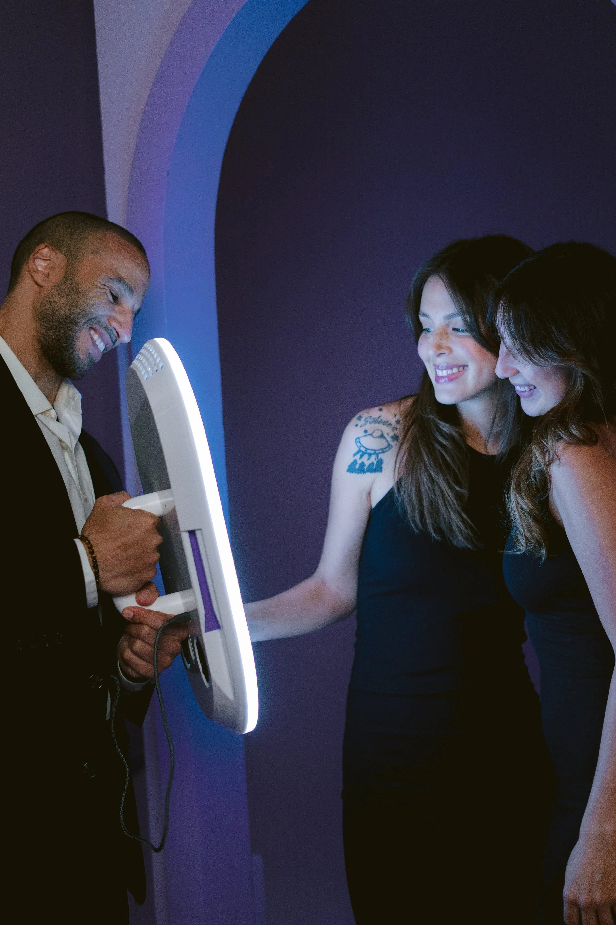 Three people smiling and interacting with a white device with a purple light, two women and one man, in a dark room with a purple background.