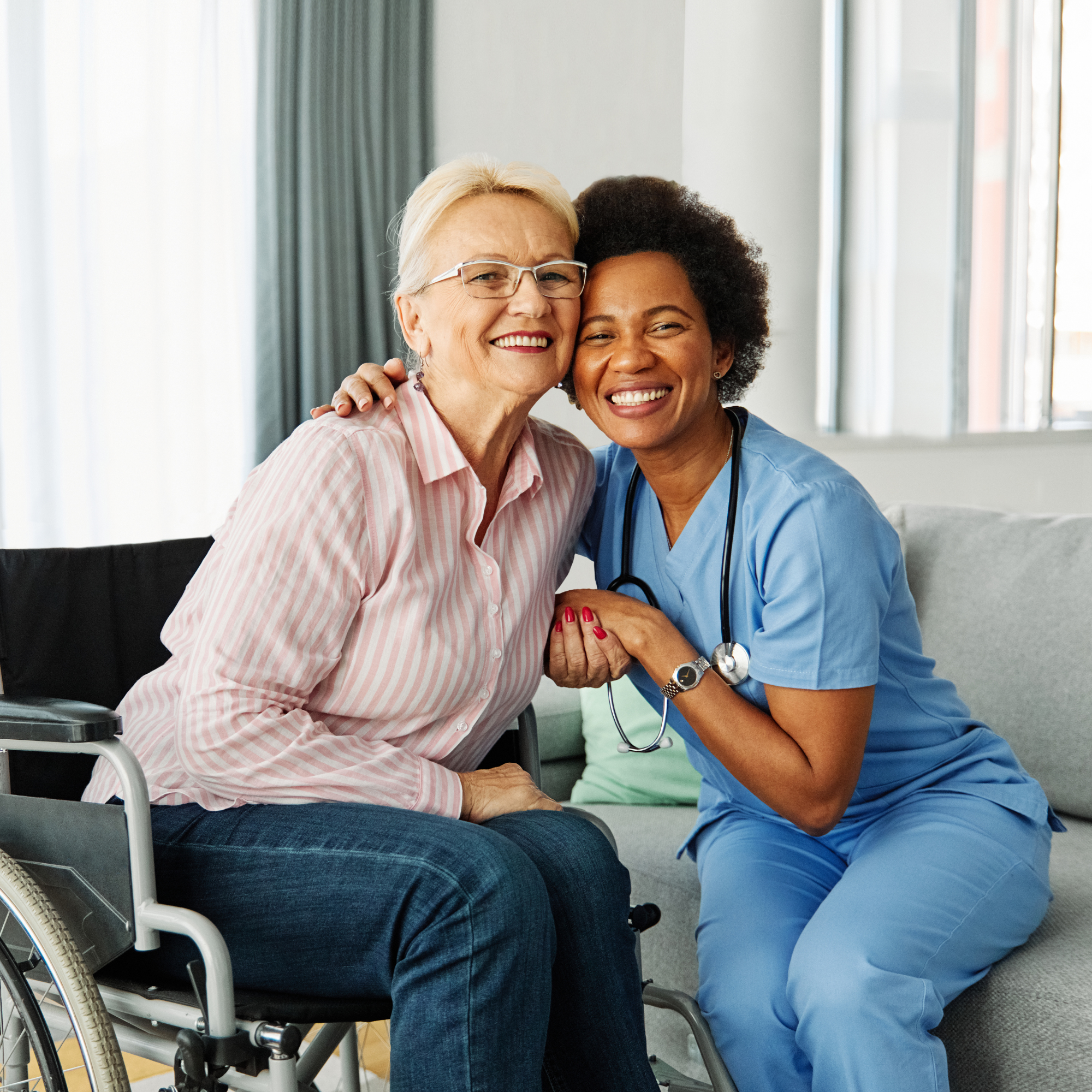A young woman in medical scrubs smiling and talking with an elderly woman wearing a green hoodie, both showing happy expressions.