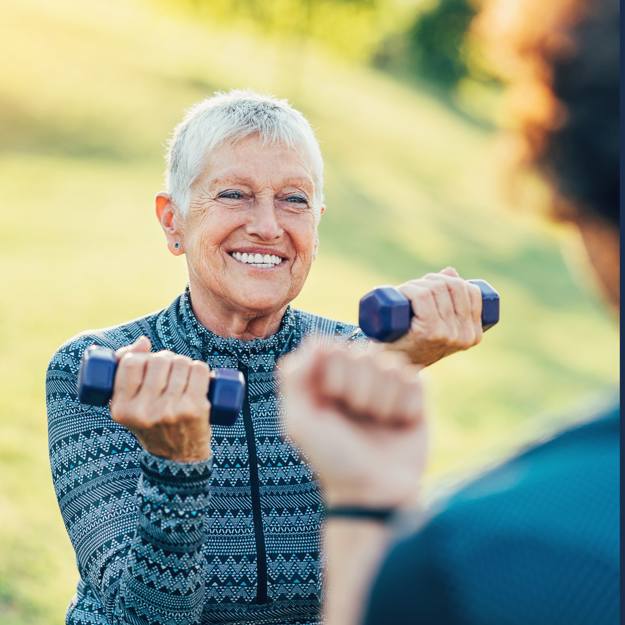An elderly woman with short gray hair smiling as she looks at someone while holding small dumbbells outdoors in a park with blurred green background.