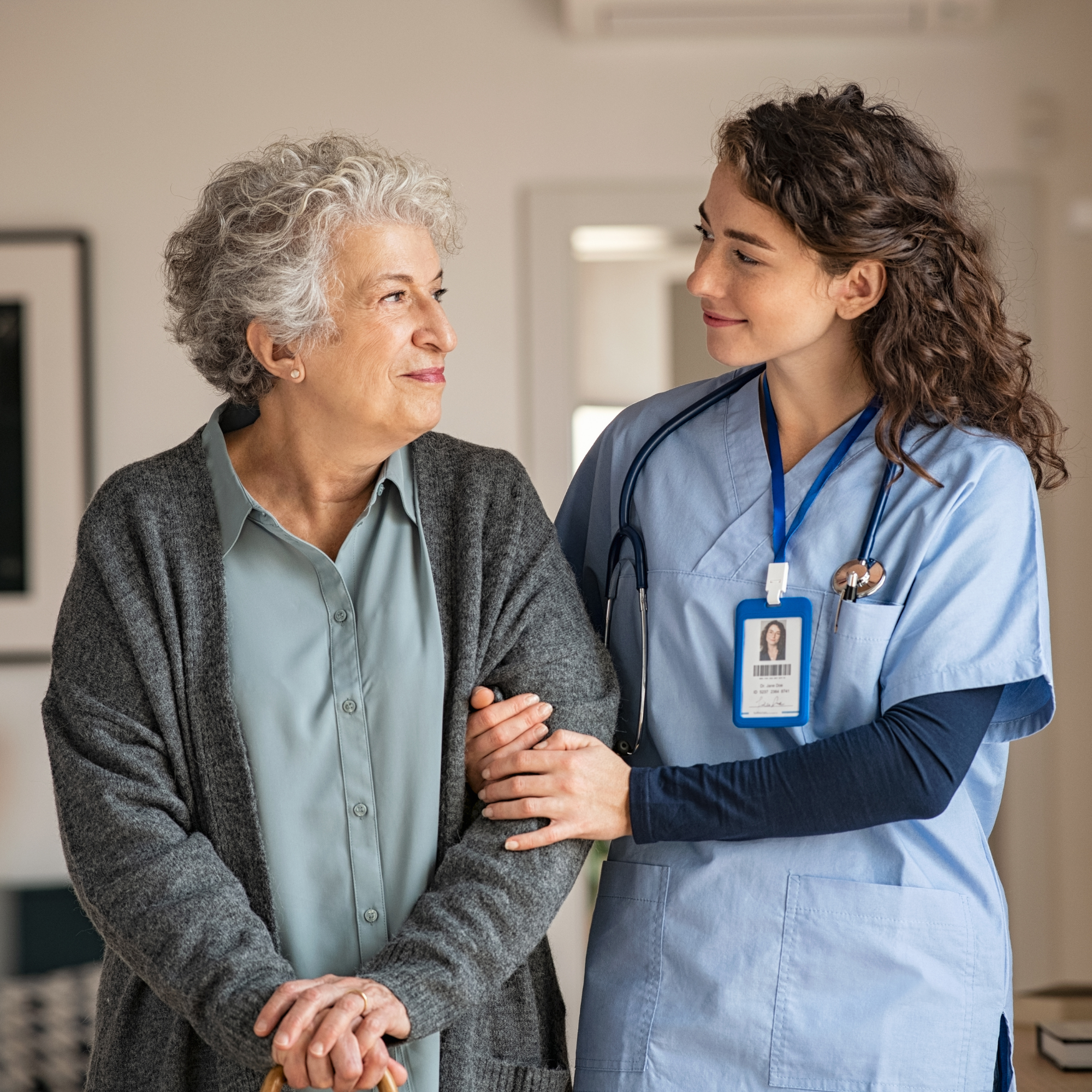 A healthcare professional, wearing scrubs and a stethoscope, gently supports an elderly woman with gray, curly hair. The elderly woman is smiling and holding a cane, and they are sharing a warm, caring moment indoors.
