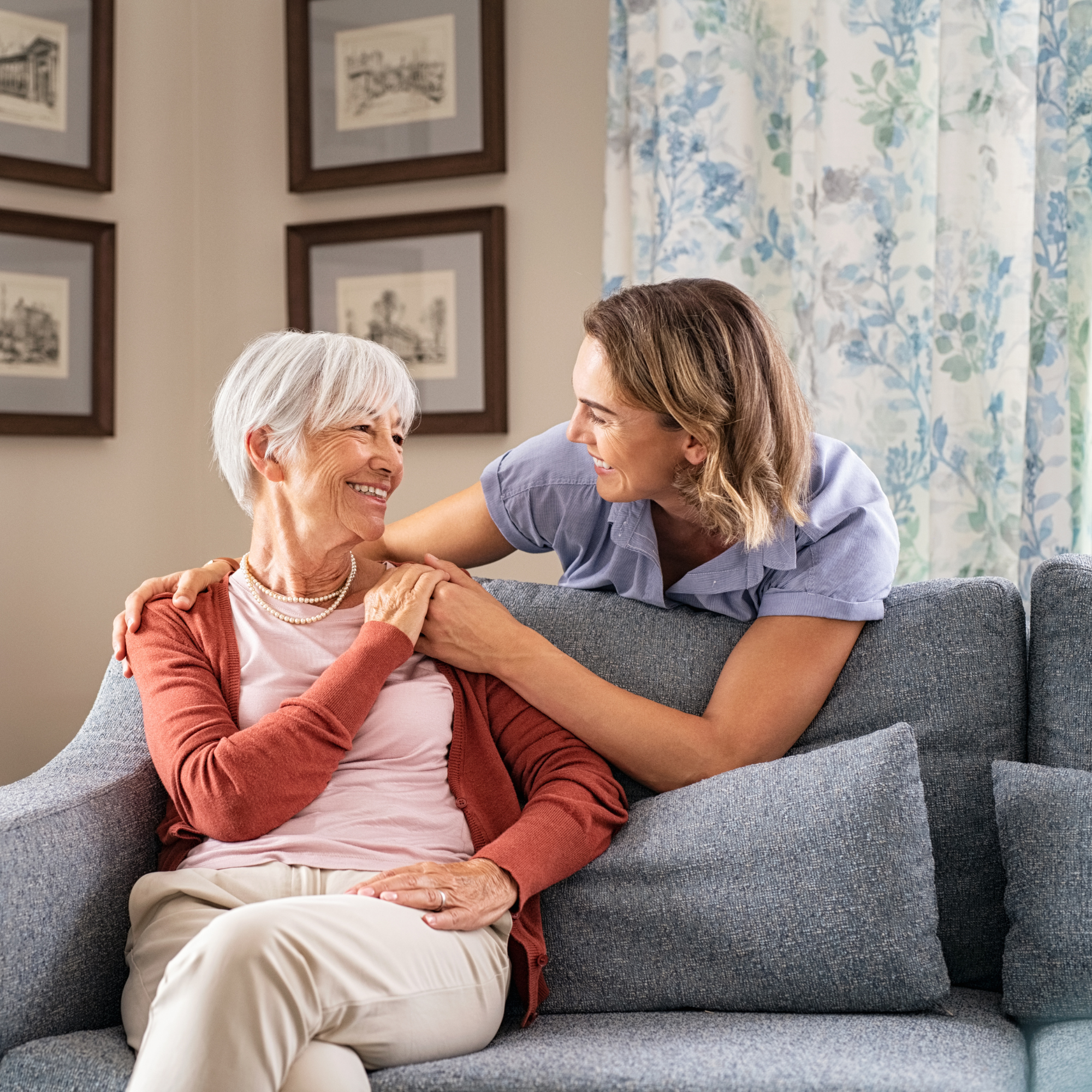 A young woman and an elderly woman are sitting on a gray couch, smiling at each other warmly. The young woman is leaning close with her hand on the elderly woman's shoulder, and they are engaging in a joyful conversation. The background includes framed artwork on the wall and floral curtains.