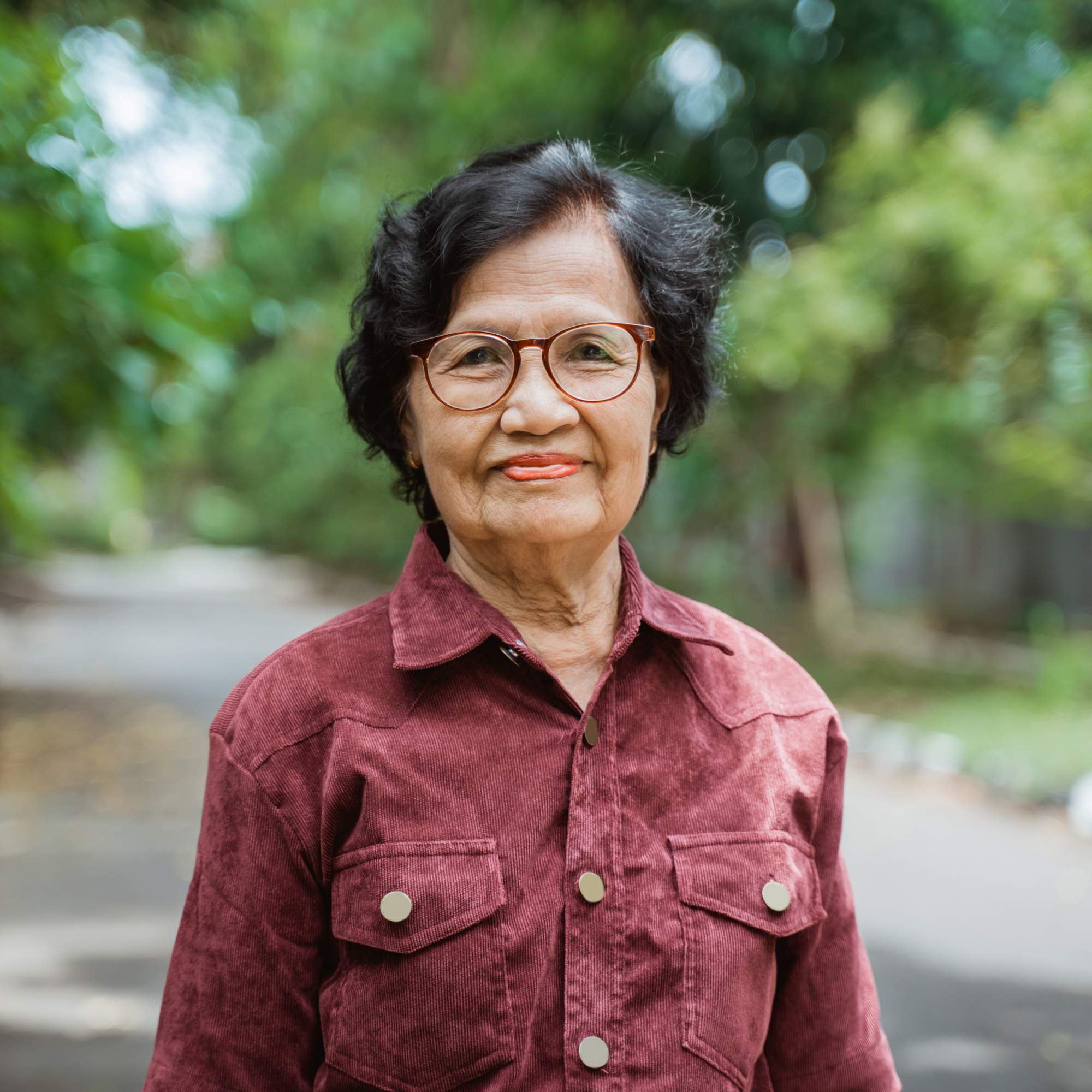 Close-up of an elderly woman with short dark hair, wearing glasses and a red jacket, standing outdoors with green trees in the background.
