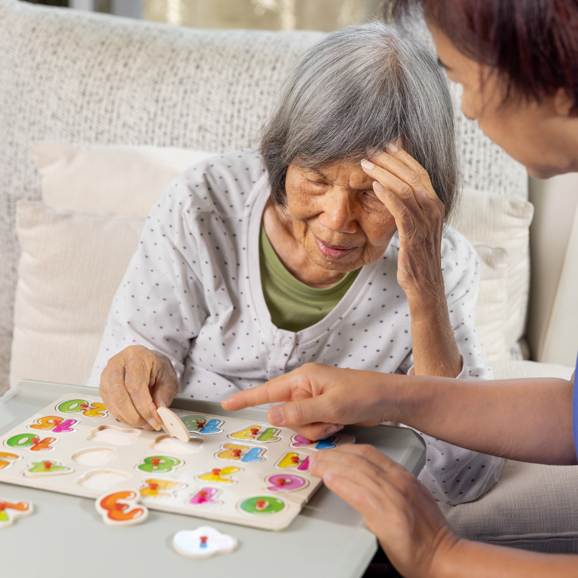A young woman showing a knitting project to two elderly women, one in a wheelchair, in a cozy, decorated room.
