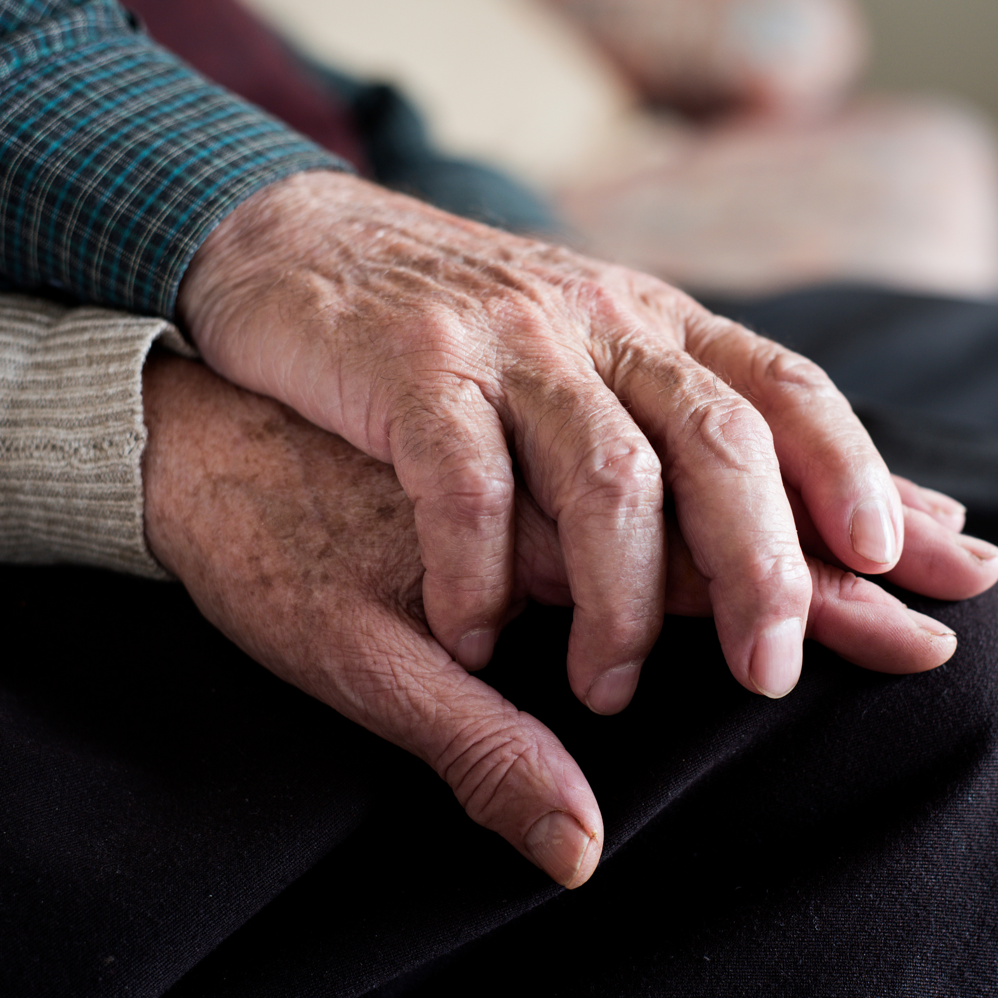 Close-up of elderly hands resting on a dark surface, with one hand gently holding the other, both showing signs of aging like wrinkles and age spots.